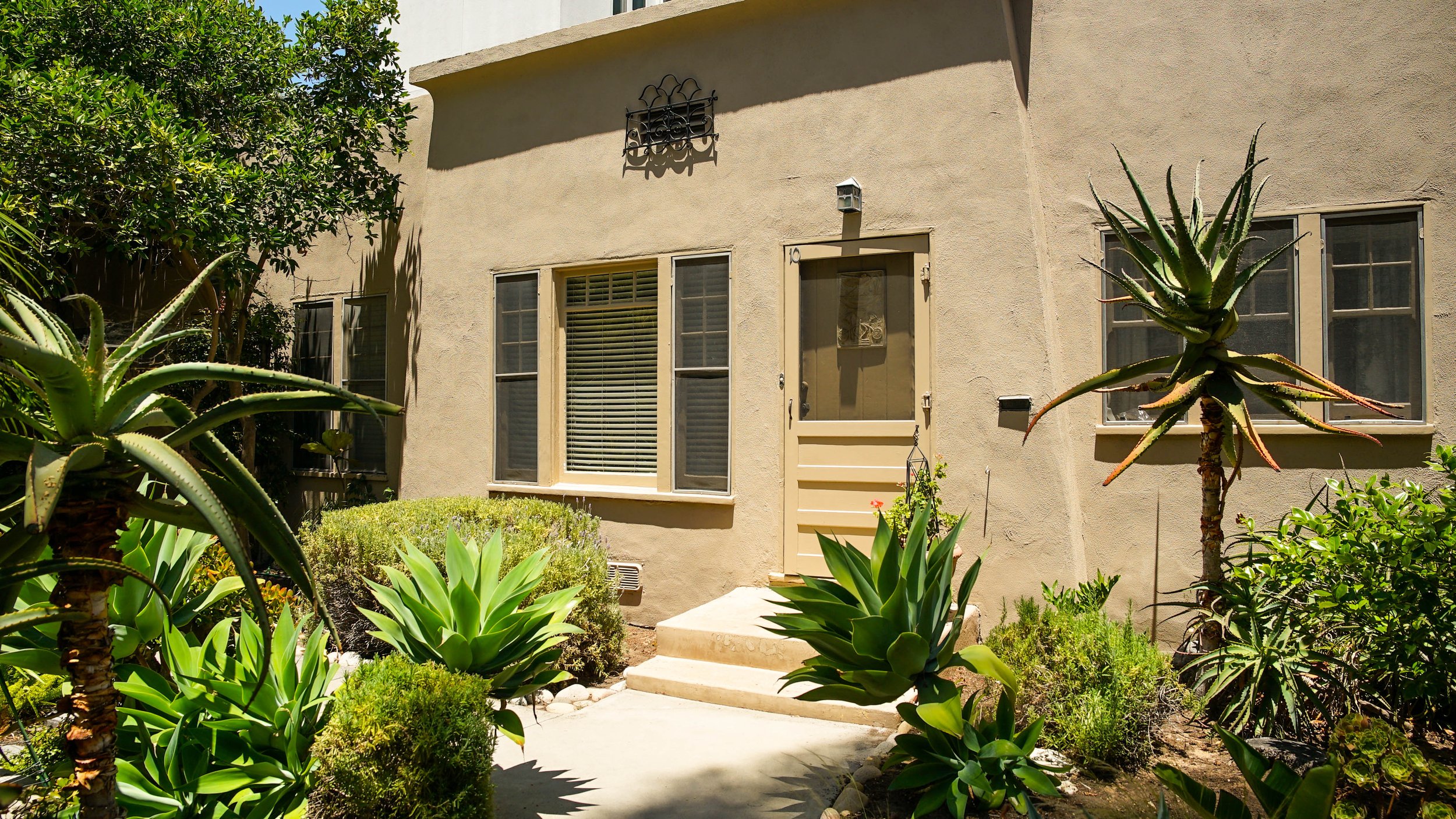 The exterior of a beige stucco house with three windows, a door, and a small porch. The yard has landscaped plants, including aloe and succulents, with sunlight casting shadows.