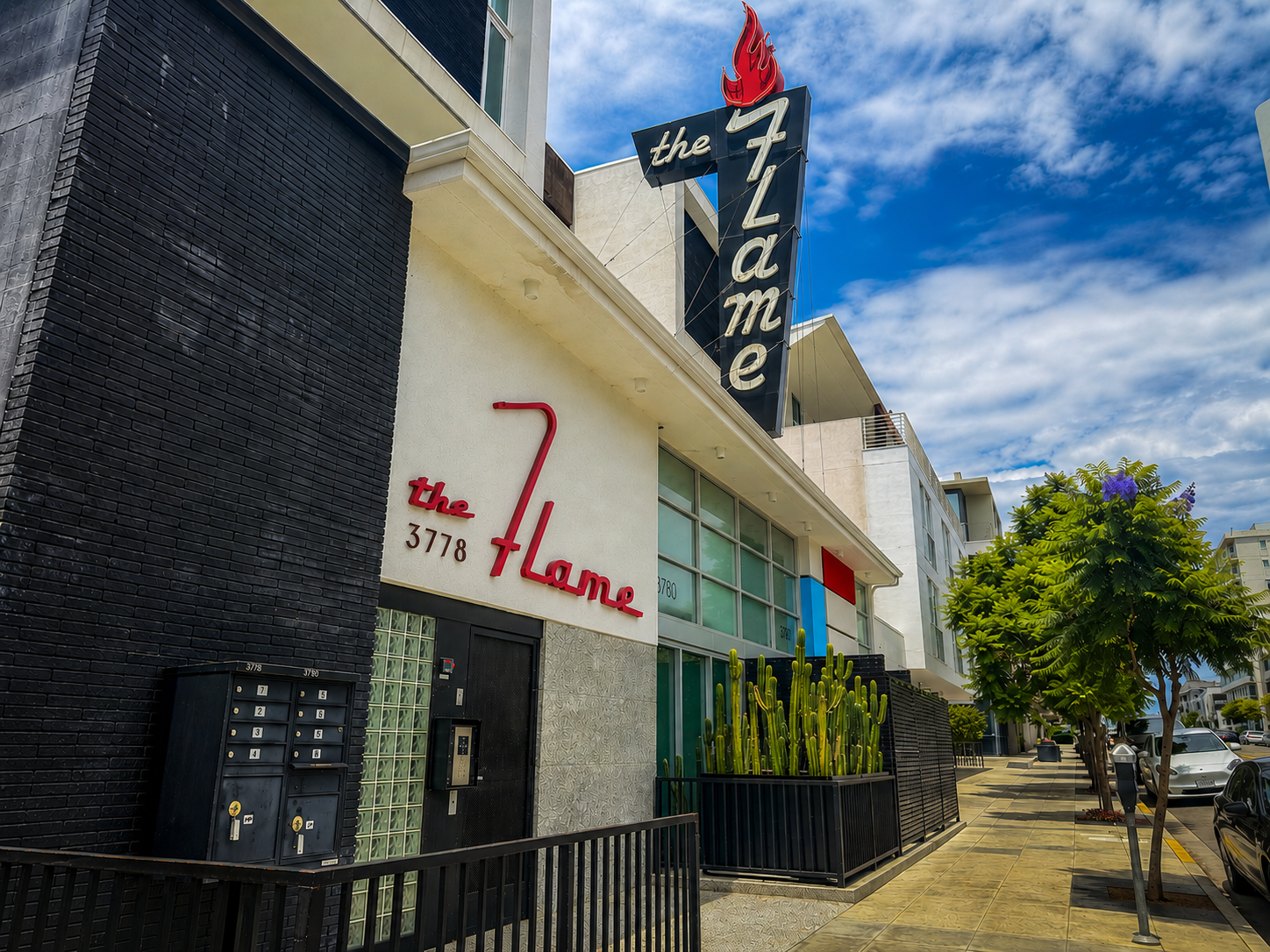 Exterior view of a modern apartment building with signs for 'the Flame' and 'the Place', cactus plants, trees, parked cars, and a partly cloudy sky.