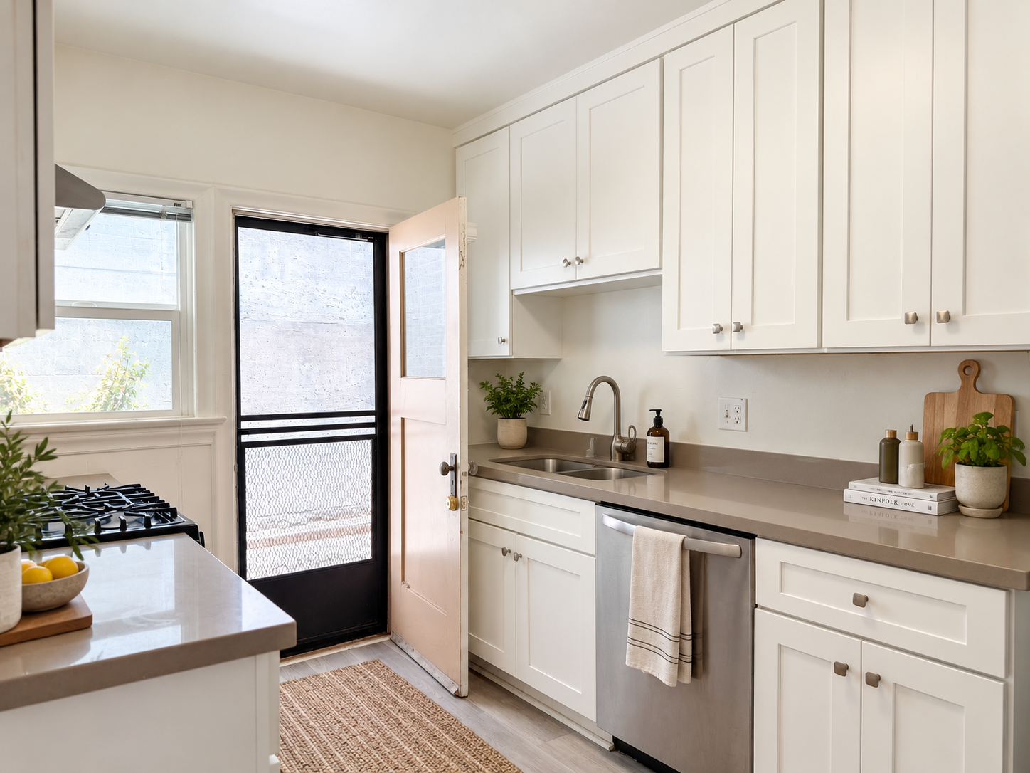 Clean kitchen with white cabinets, beige countertop, stainless steel sink, potted plants, soap dispenser, books, and a door leading outside with a small window, natural light, and a woven rug on the floor.