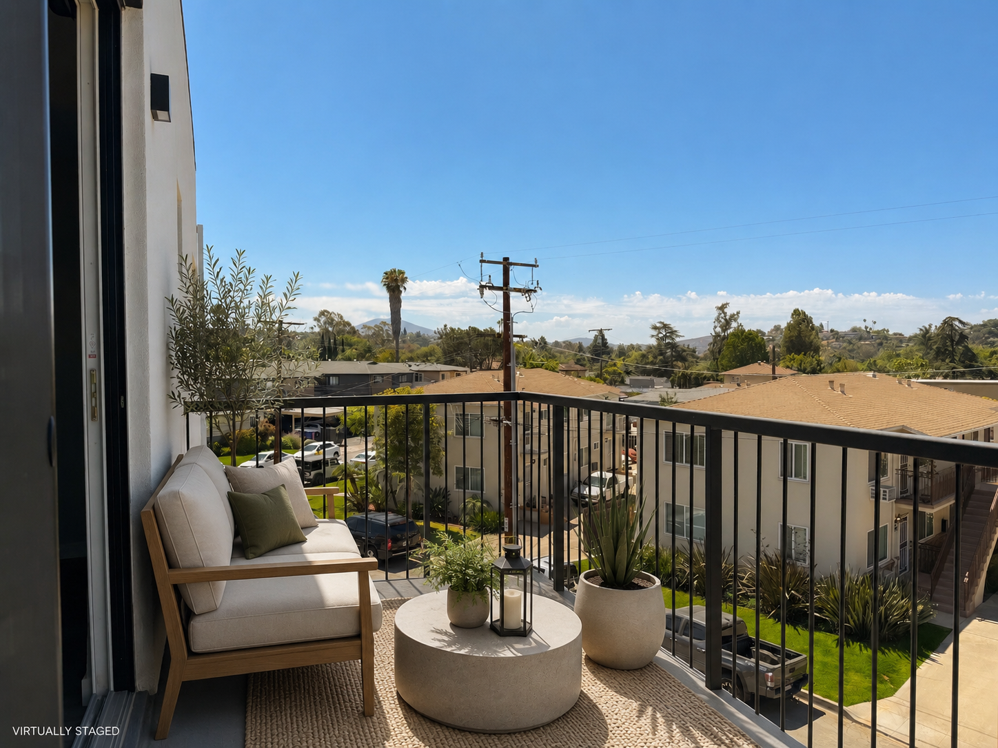 Balcony with beige sofa, potted plants, and a lantern, overlooking a residential neighborhood with rooftops, trees, and utility poles under a clear blue sky.