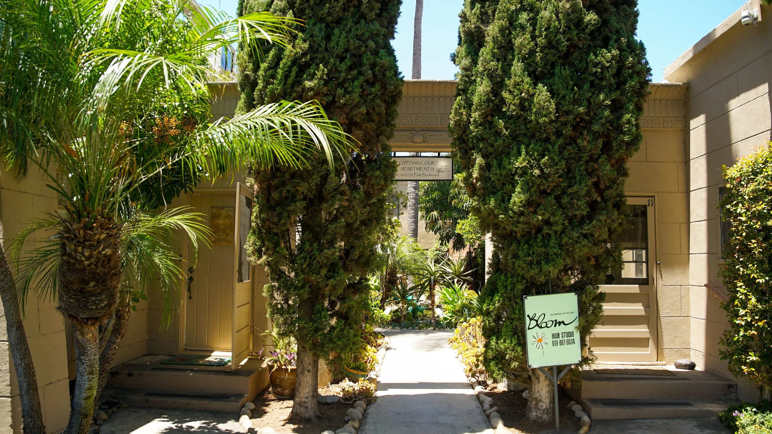 Entrance to a courtyard with lush tropical plants and tall trees, leading to apartment buildings named 'Egyptian Court Apartments.' A sign for a hair studio is visible near the stairs on the right.