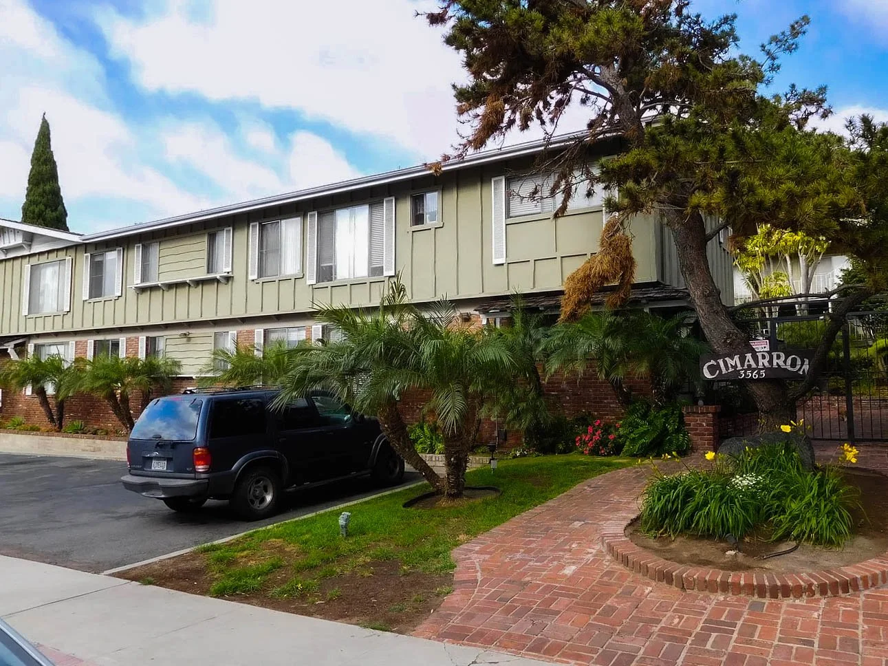 A multi-unit residential building with green siding, brick foundation, and multiple windows, surrounded by landscaped plants and trees, with a driveway and parked black SUV in front. A sign near a large tree reads "CIMARRON 3565."