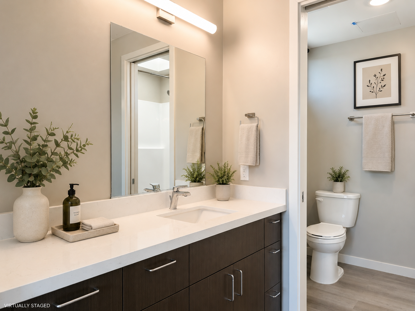 Modern bathroom with a large mirror above a white quartz countertop with a sink and plants, and a toilet in an adjacent room with wall art and towels.