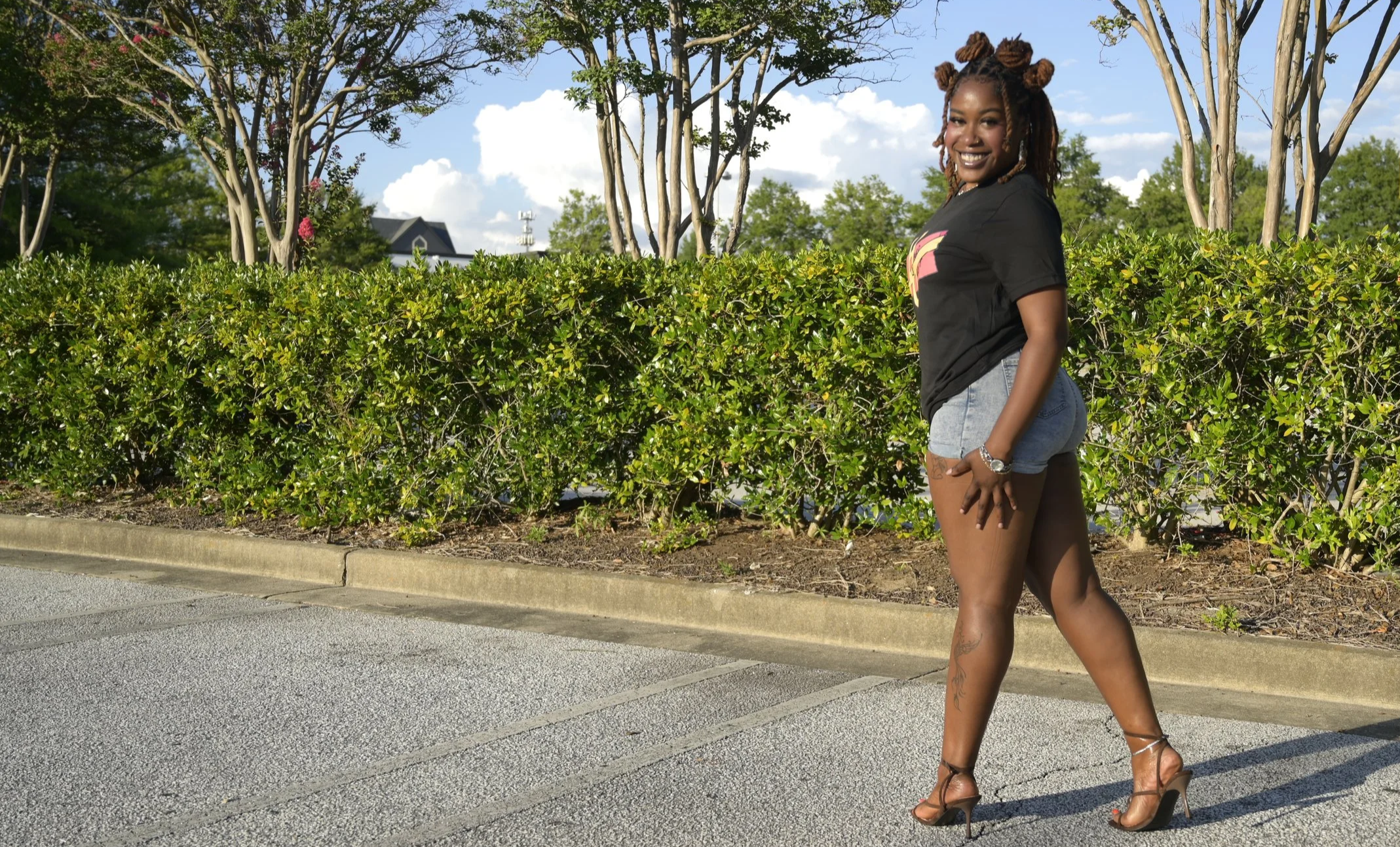 Aura Laura, host of Hard & Uncut, smiling over her shoulder while walking outdoors in a black graphic tee, denim shorts, and heeled sandals.