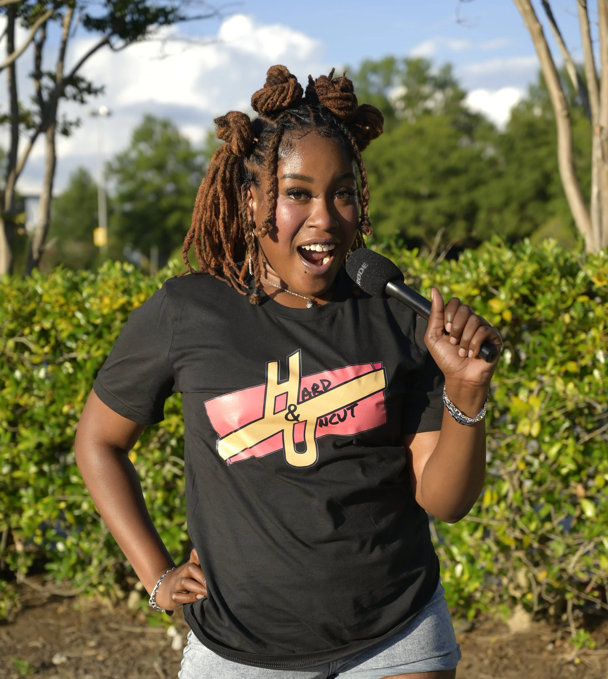 Aura Laura, host of Hard & Uncut, smiling with a microphone to her face while outdoors in a black graphic tee, and denim shorts