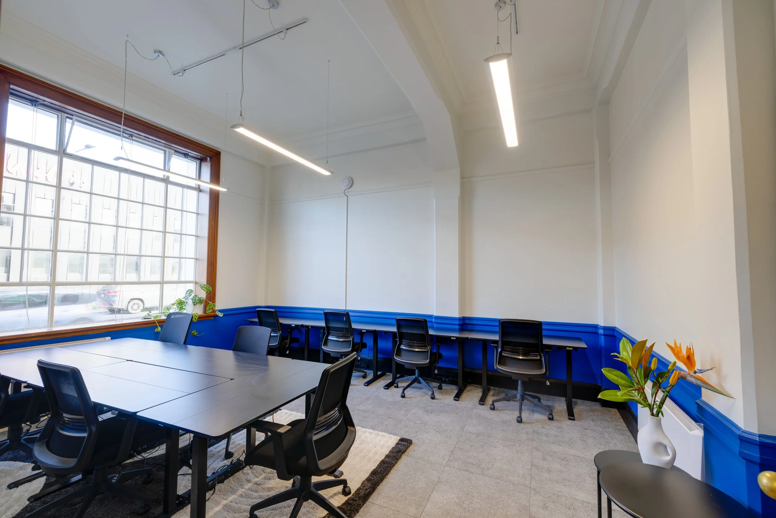 A private office space with a large window, black chairs around a black table, a row of chairs with a blue trim along the wall, and a white vase with orange flowers on a small table in Bad Company Hawkes Bay. 
