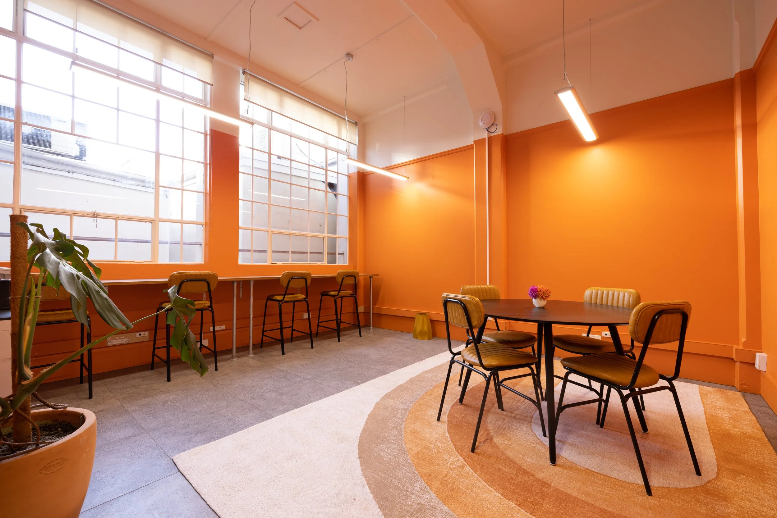 Private office space within Bad Company Hawkes Bay, with large windows, a round wooden table with four chairs, a small vase with purple flowers, a rug, and a row of bar stools along the window. 