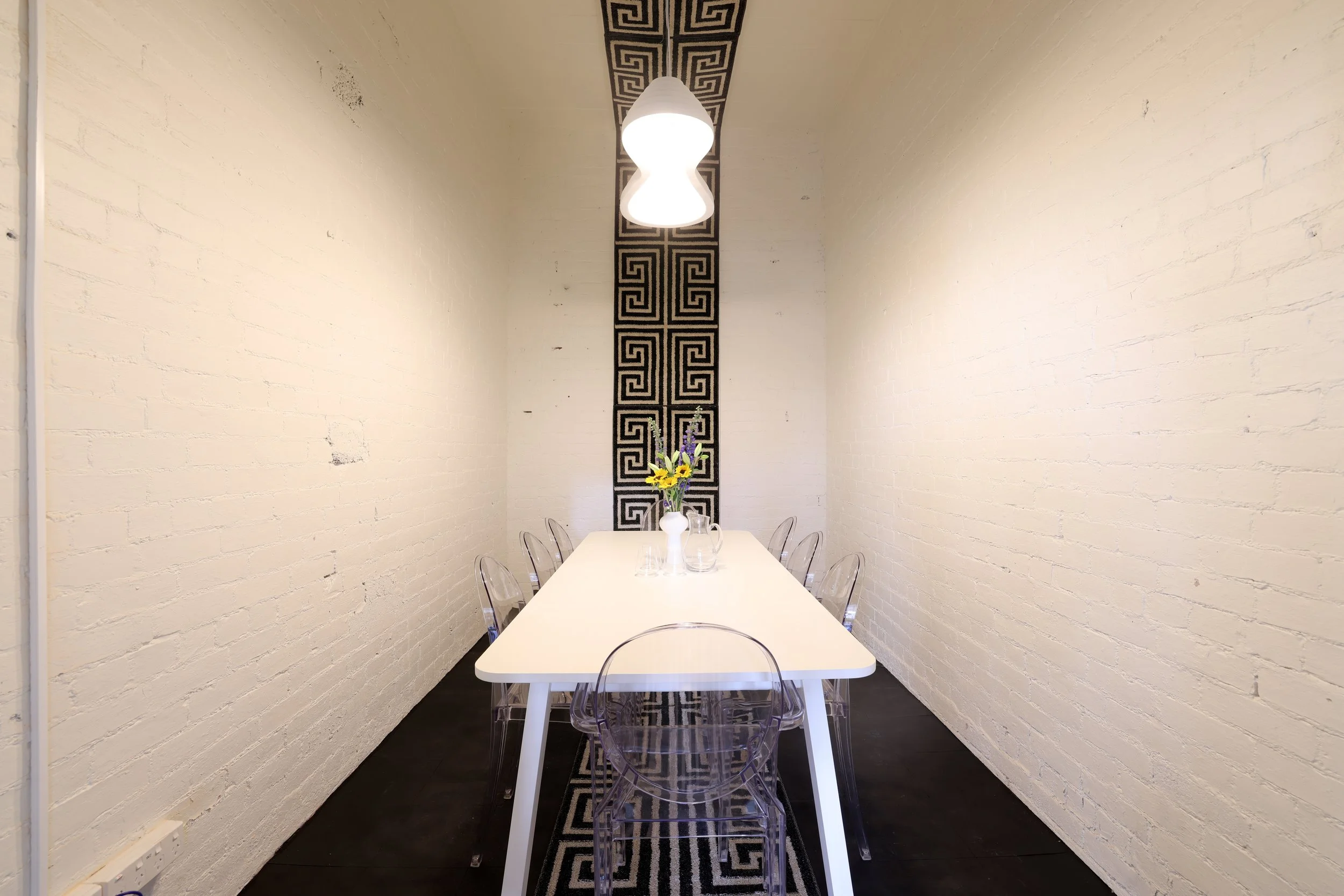A 8 person board room, bookable for meetings with a long white table, and transparent chairs, decorated with a vase of flowers. There is geometric black wallpaper strip and hanging light fixture Bad Company Hawkes Bay, a co-working space, Hastings. 