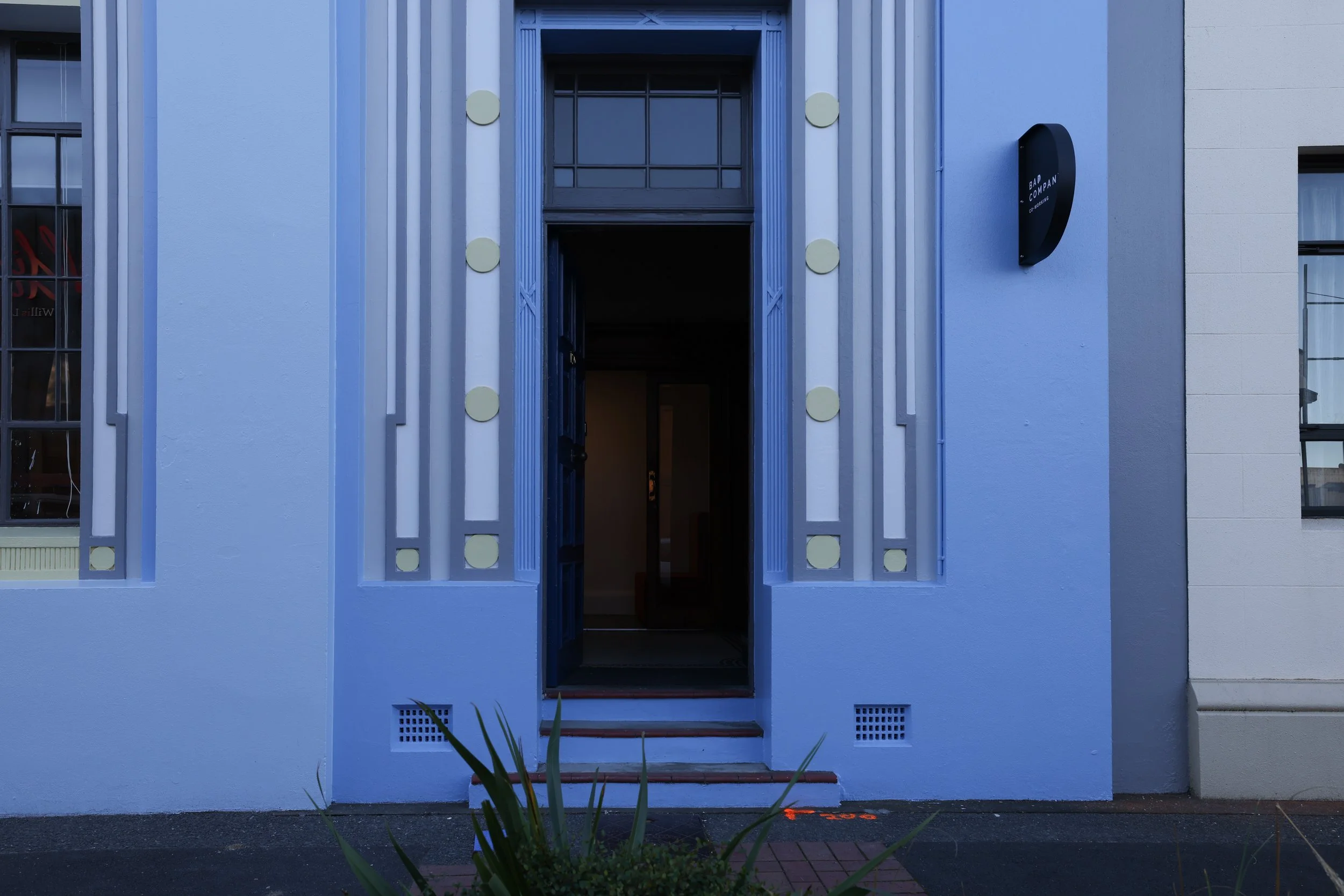 The exterior of a modern blue building with a sign that reads ‘Bad Company Co-Working’ on a black and blue circular logo. This is the front door of a great co-working space in Hastings, New Zealand. 
