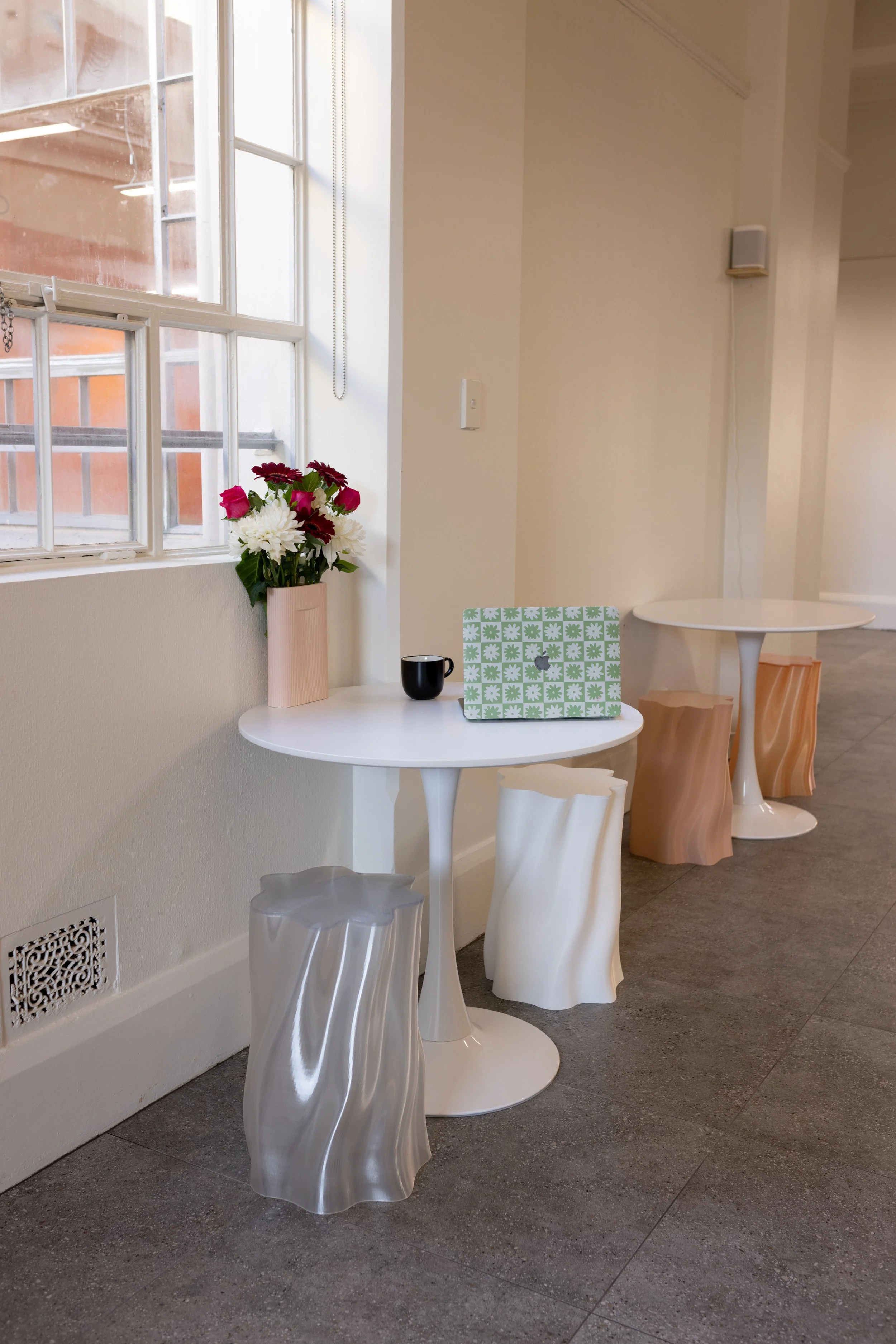 Interior scene with two white round tables, one decorated with pink and white flowers, a black cup, and a decorative box, set in front of a window with orange and brown outside views. Bad Company Hawkes Bay, a co-working space based in Hastings, NZ