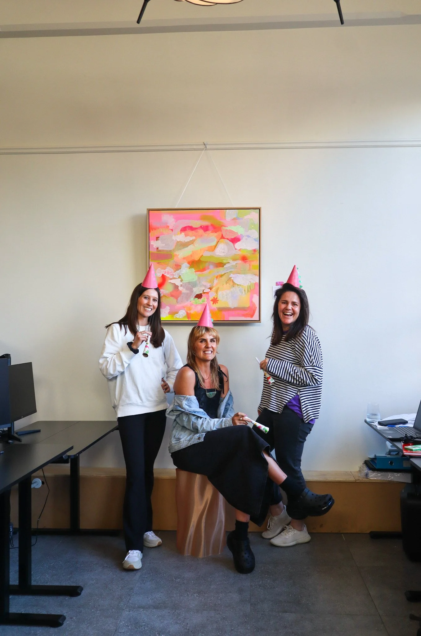 Three women celebrating at a party, wearing pink party hats, with colorful artwork on the wall behind them. They are celebrating at Bad Company Hawkes Bay. A co-working space in Hastings New Zealand, that likes to celebrate. 