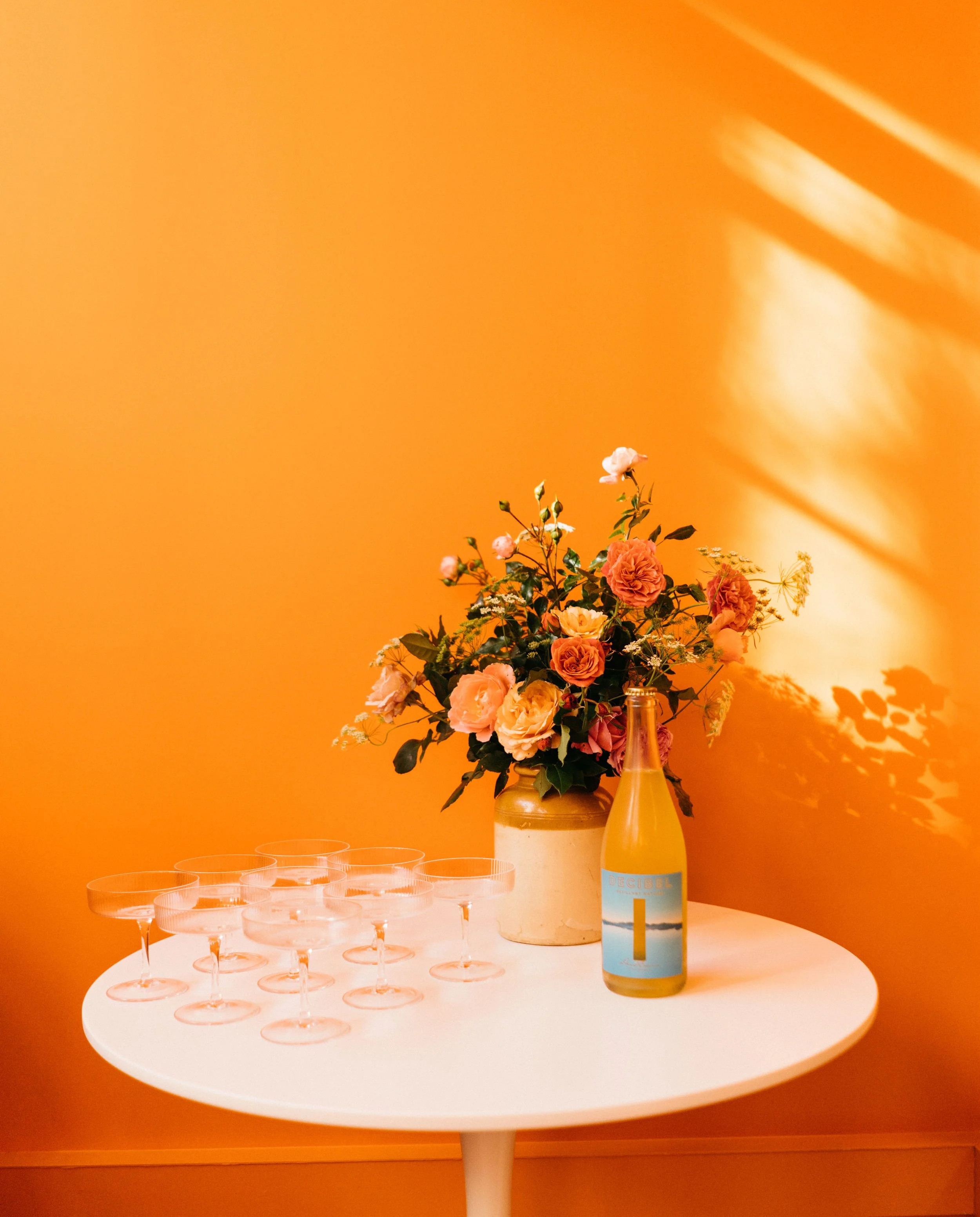 A white round table with a flower arrangement of peach and pink roses in a beige vase, a bottle with a blue label, and several empty glasses against an orange wall with sunlight shadows at Bad Company Hawkes Bay, a co-working space