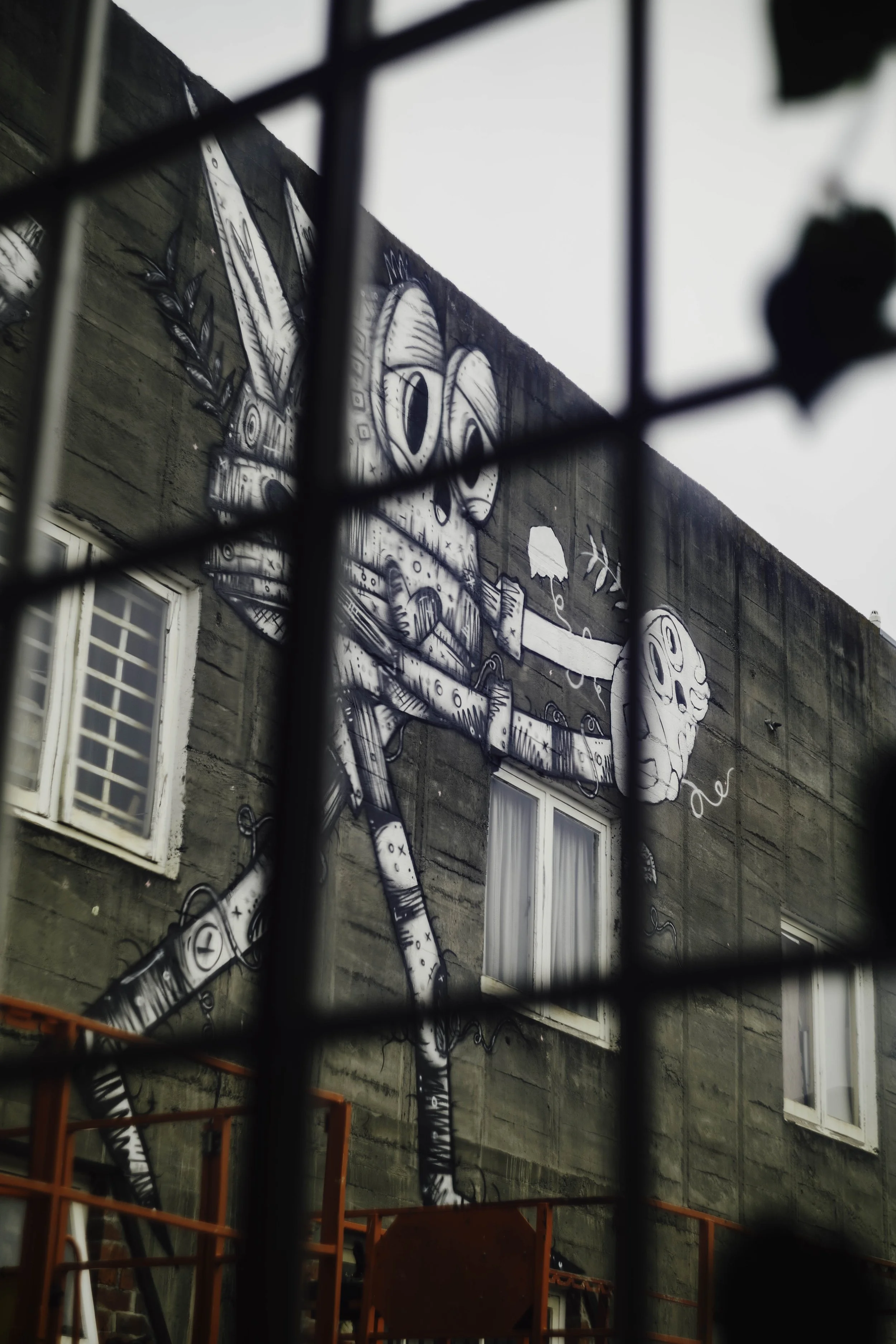 Street art mural of a robot with feathers, holding a mask and a drawing tool, painted on a building wall, seen through a black metal fence at Bad Company Hawkes Bay, a co-working space based in Hastings, New Zealand.