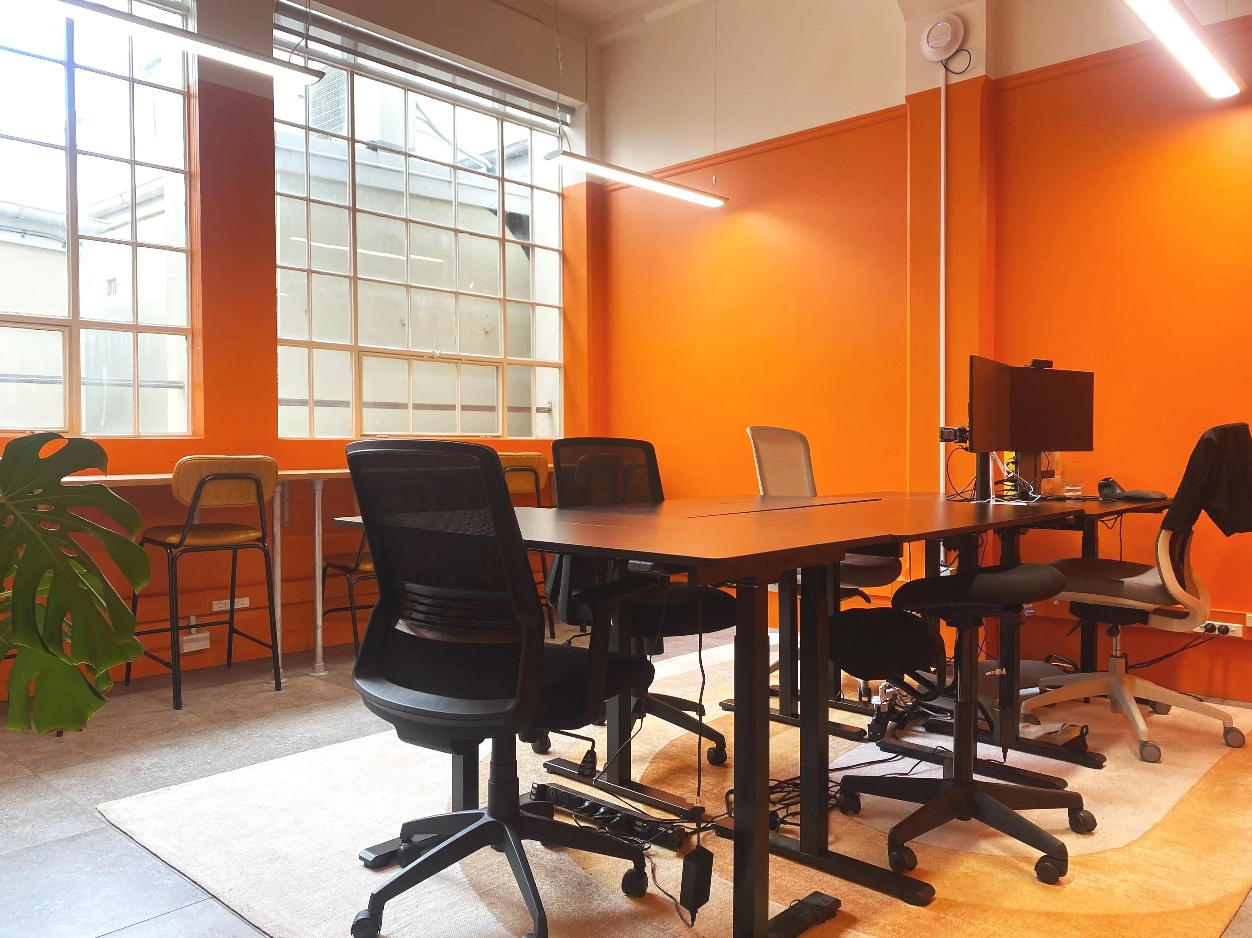 A modern private office conference room with orange walls and large windows. Contains a black conference table surrounded by office chairs. Includes a small desk with a computer monitor. Based within Bad Company Co-working space in Hawkes Bay. 