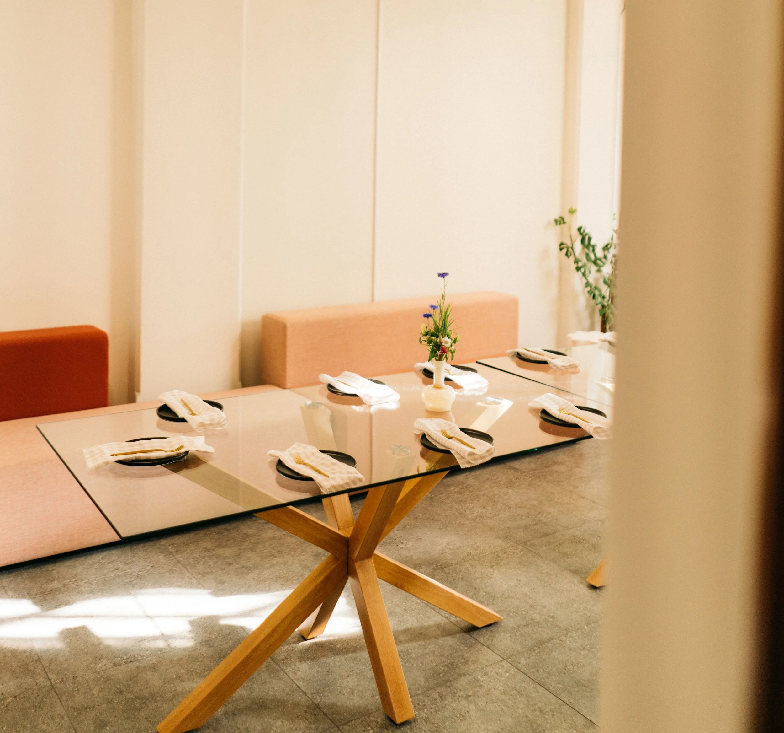 Dining table set with six places, each with a black plate, white napkin, and cutlery, and a small vase with flowers in the center, in a well-lit room with beige walls.