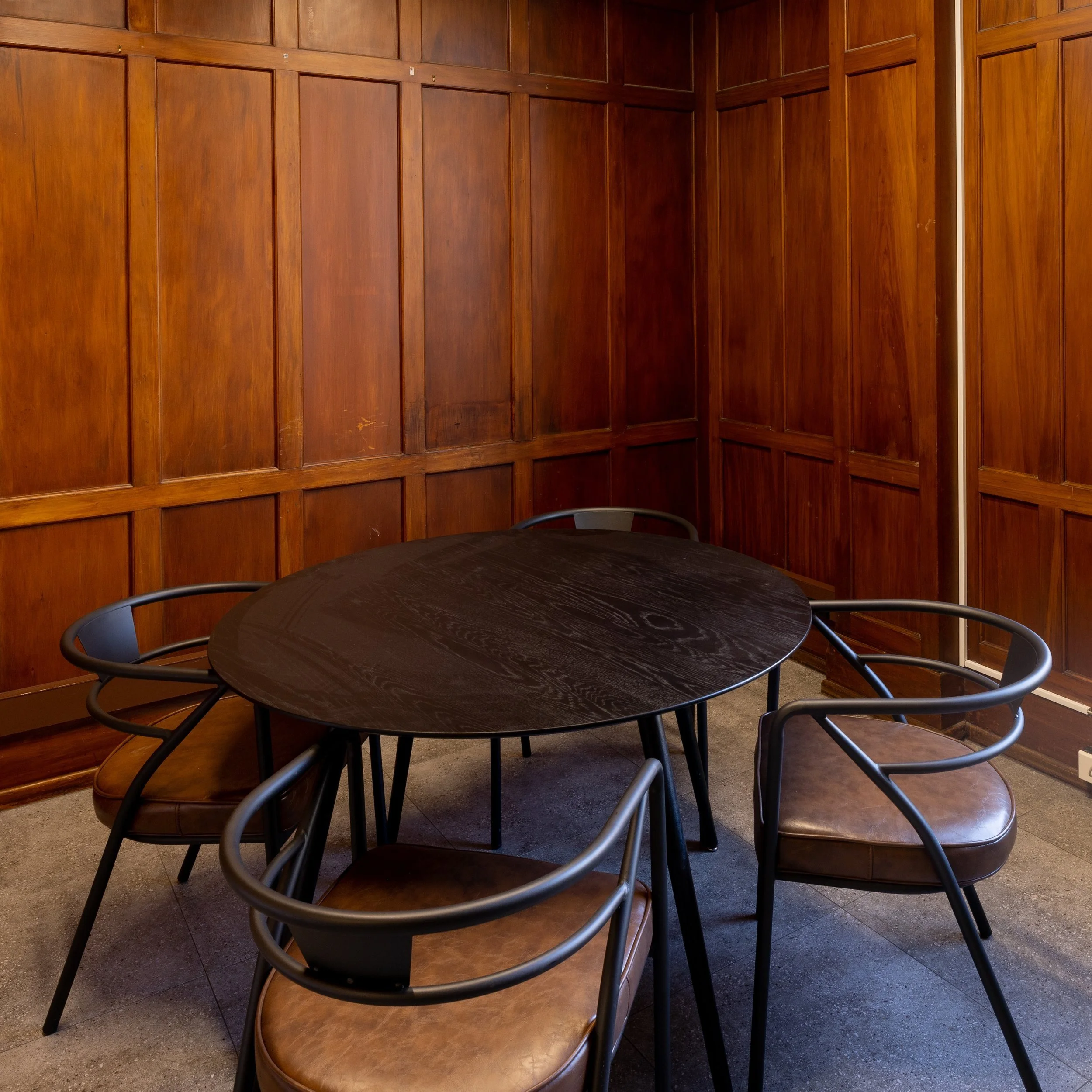 A small round black table with three brown cushioned chairs facing wood-paneled walls in a cozy room.