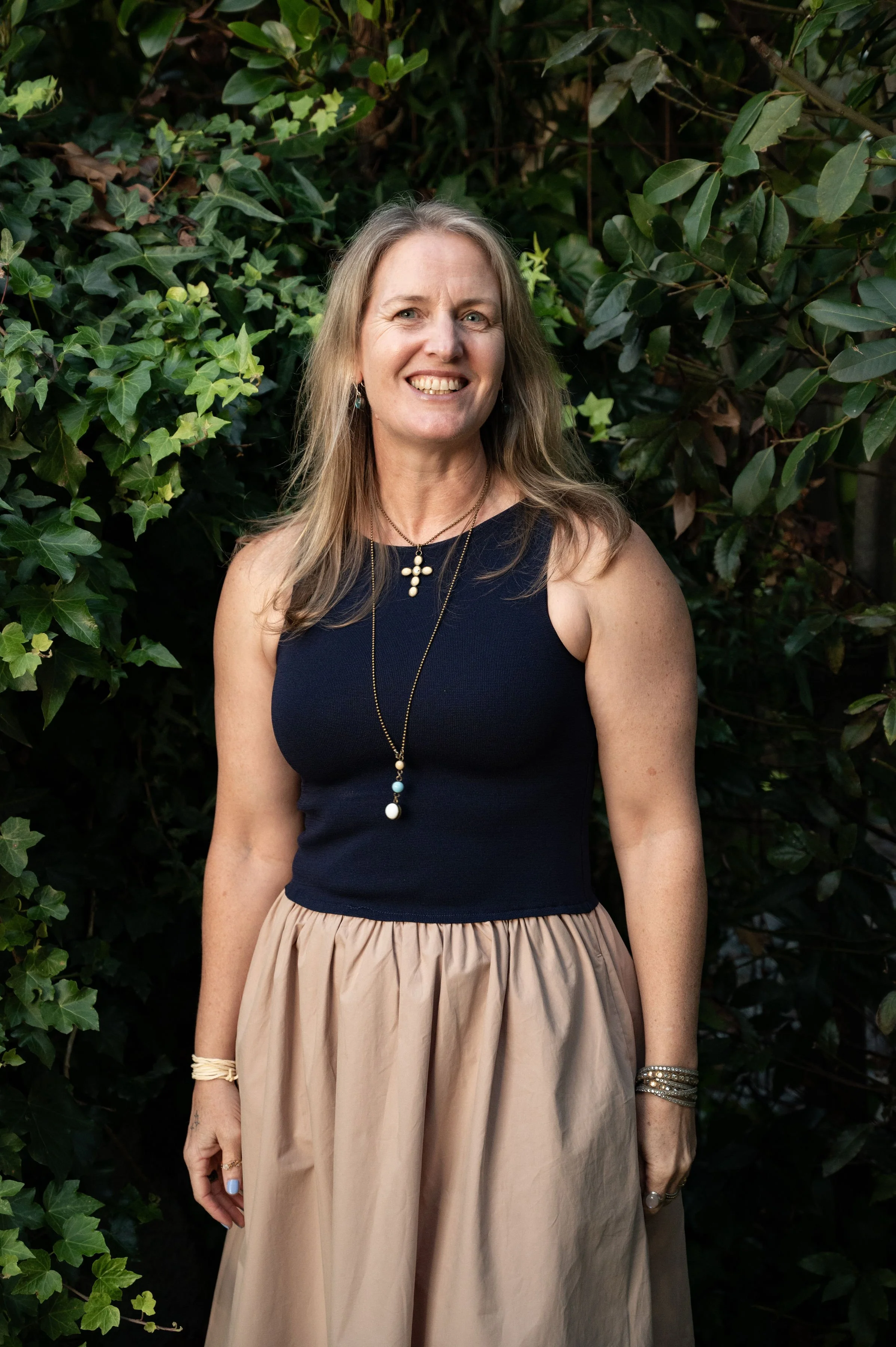 A woman with long blonde hair, smiling, wearing a navy sleeveless top, beige skirt, and jewelry, standing outdoors in front of green leafy bushes.