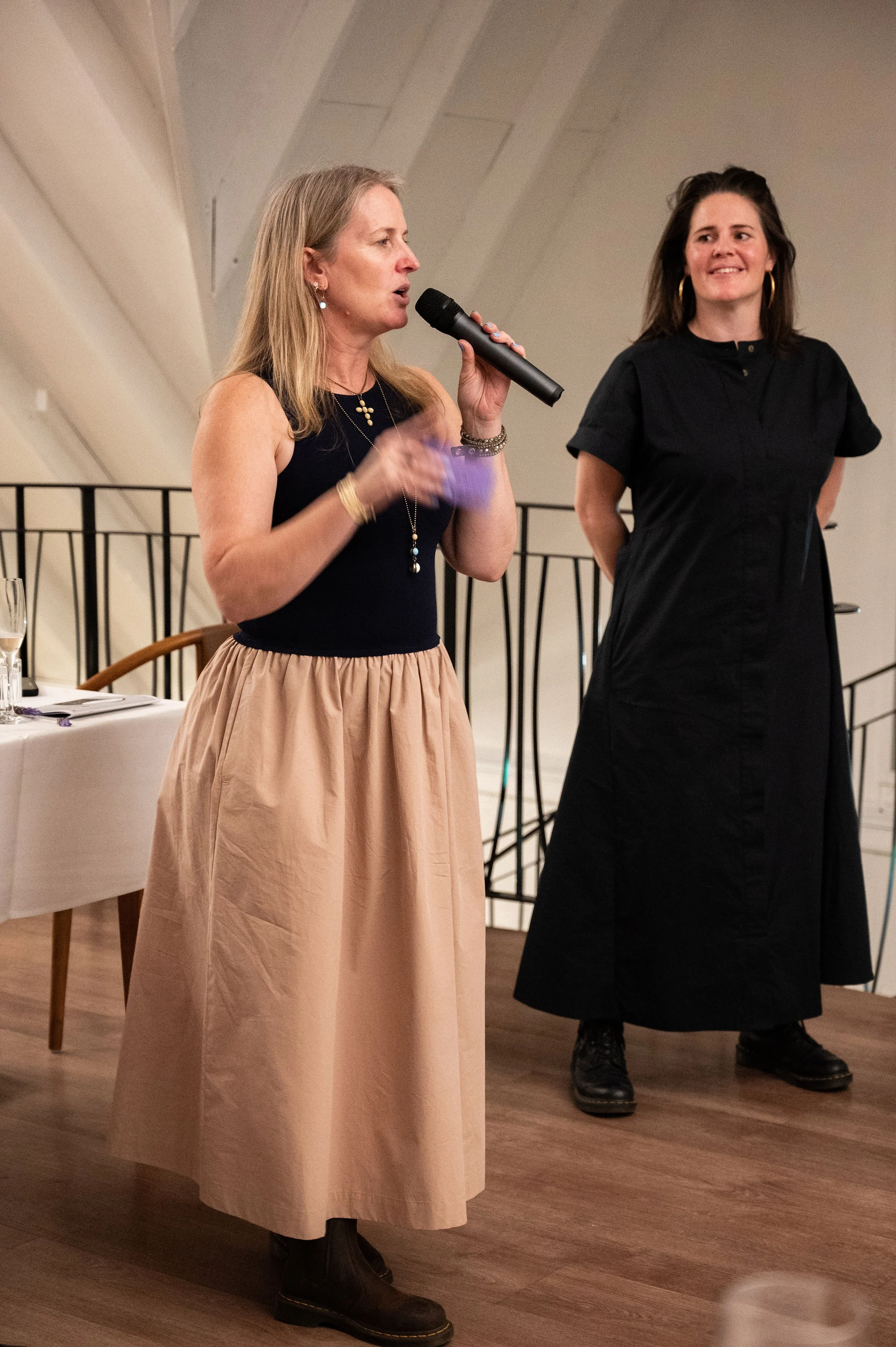 A woman in a black sleeveless top and beige skirt is speaking into a microphone during an event, with another woman in a black dress standing behind her.