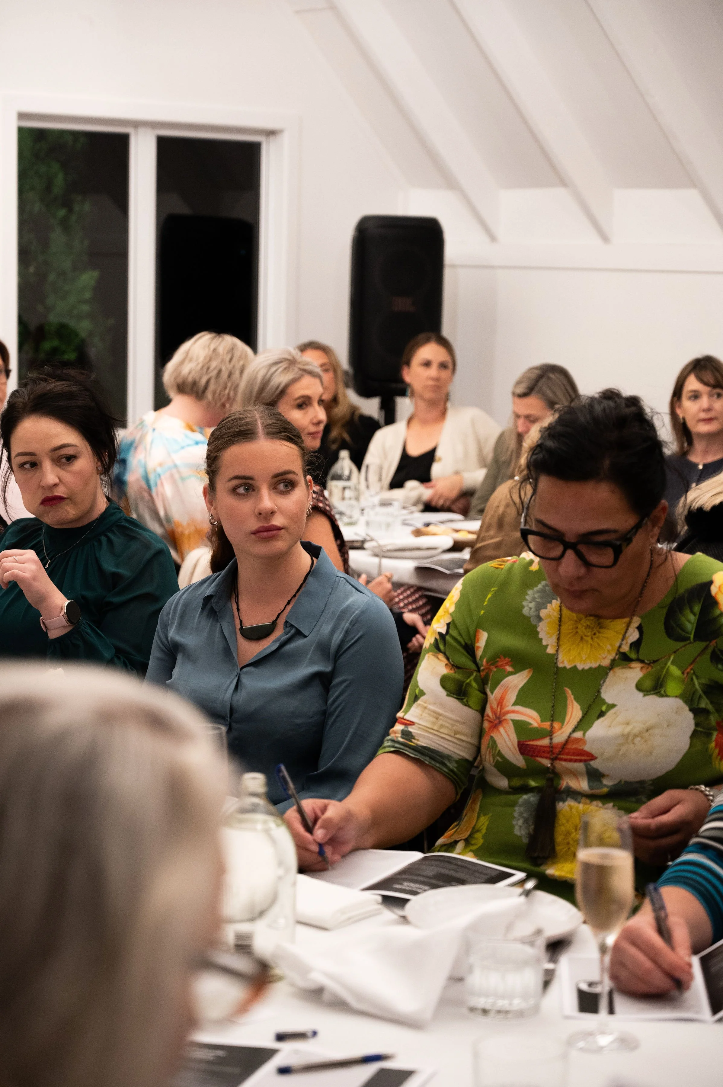 A group of women seated at a table during a meeting or conference in a well-lit room, with some taking notes and others listening attentively.