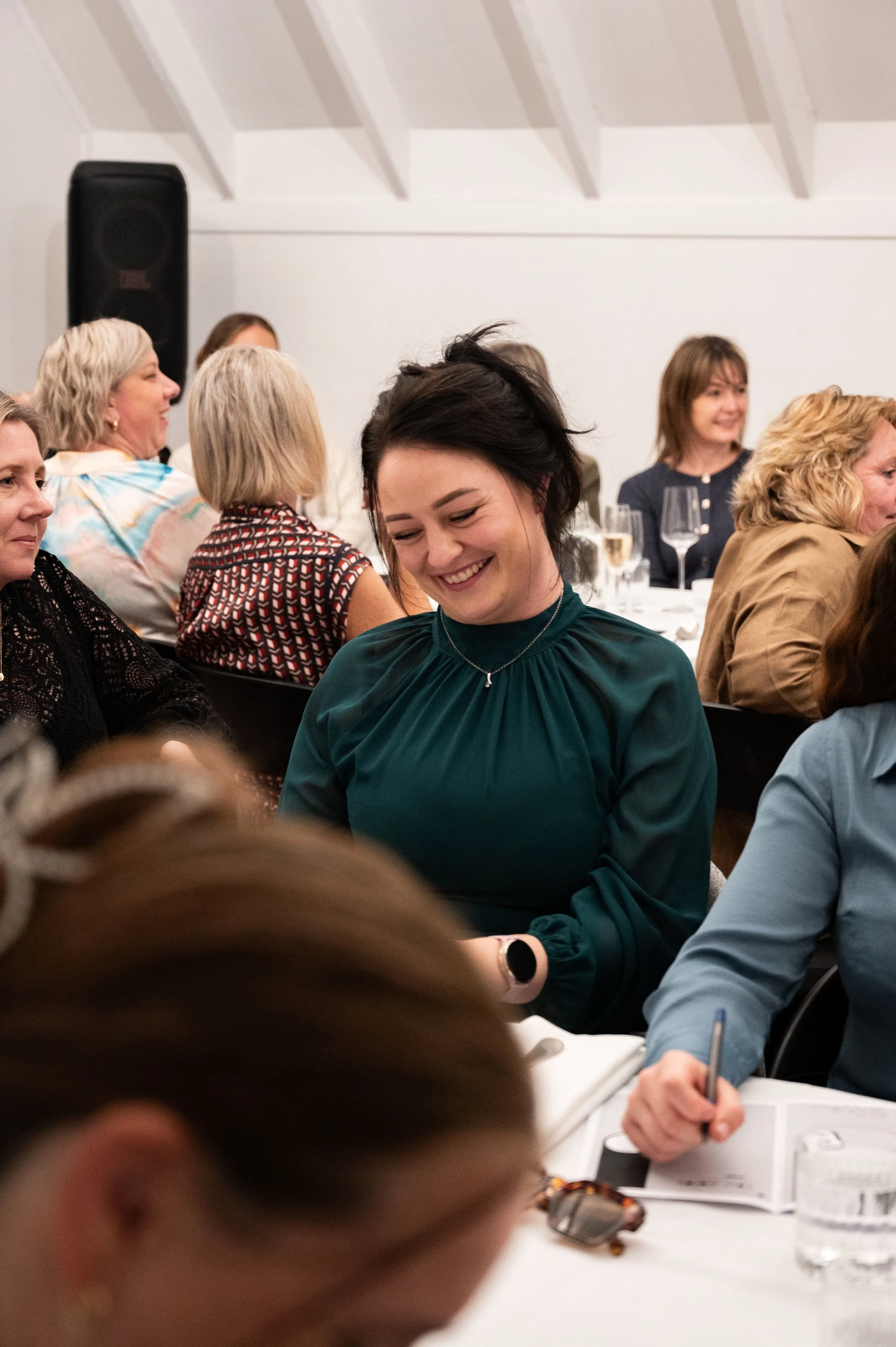 A group of women sitting at a dinner table, smiling and enjoying conversation in a well-lit, indoor setting.