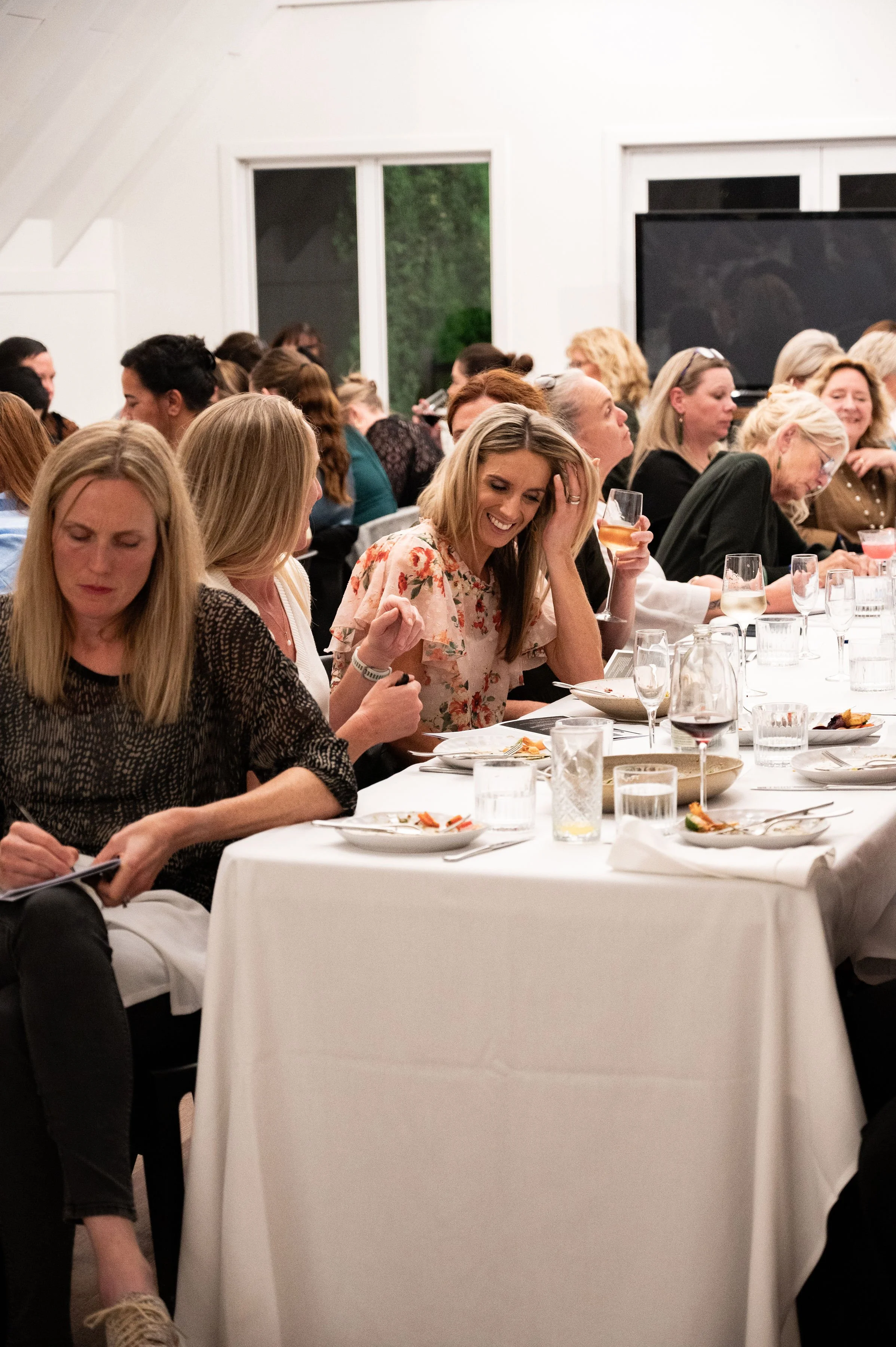 A group of women sitting at a long table during a social event or dinner, laughing and engaging with each other, with plates, glasses, and beverages on the table.