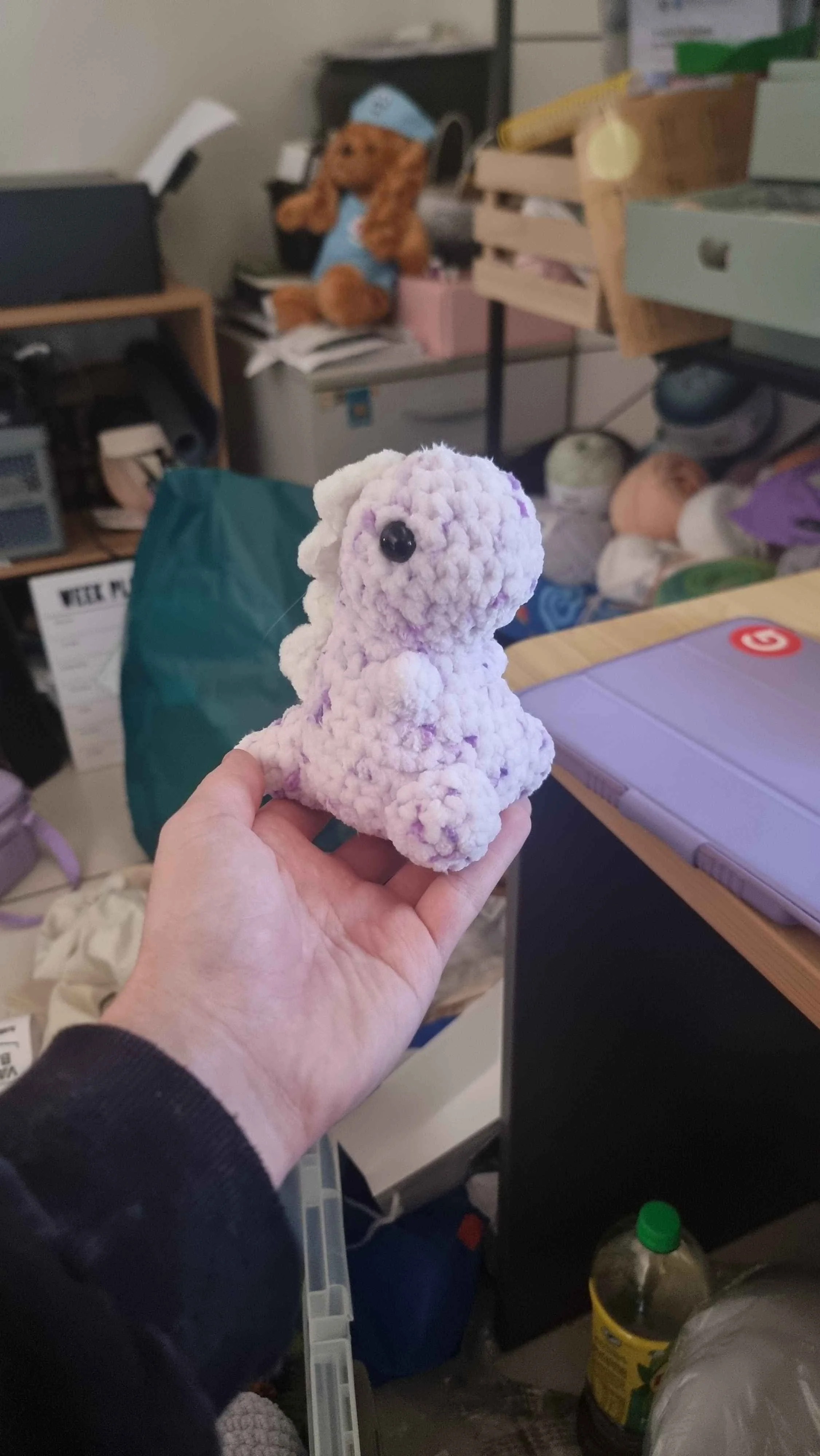 Hand holding a plush toy resembling a alpaca or llama in a cluttered room with shelves, a teddy bear, and various objects.