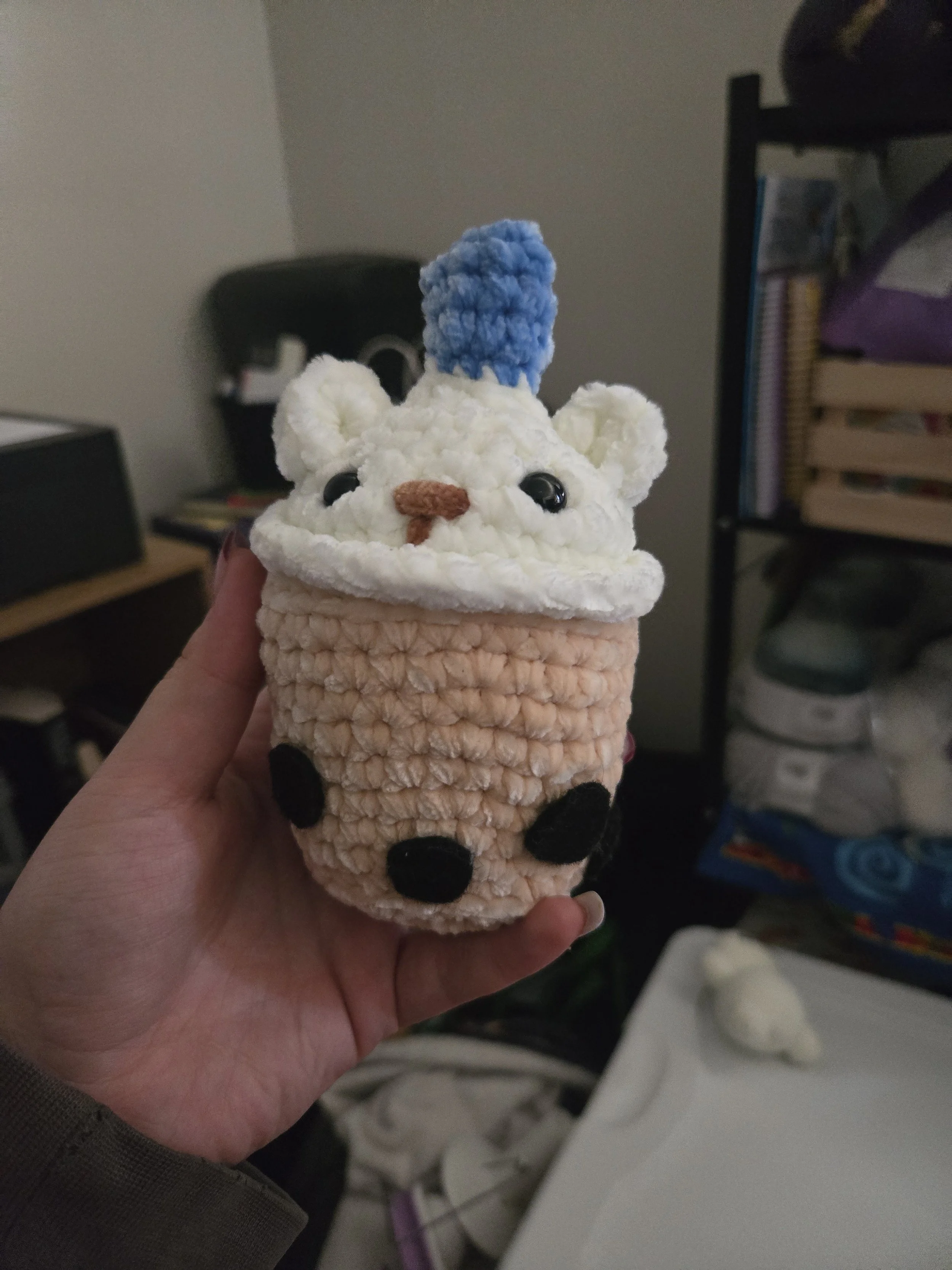 A plush teddy bear with a blue pom-pom on its head, held in a person's hand.