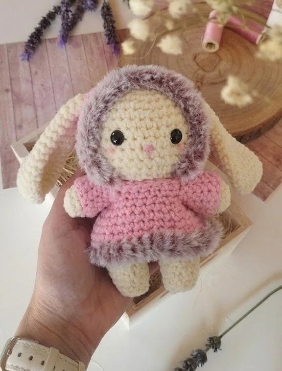 A crocheted plush toy of a cute bunny with large black eyes, a small pink nose, and long floppy ears. The bunny is wearing a pink dress with gray furry trim, and is being held in a person's hand.