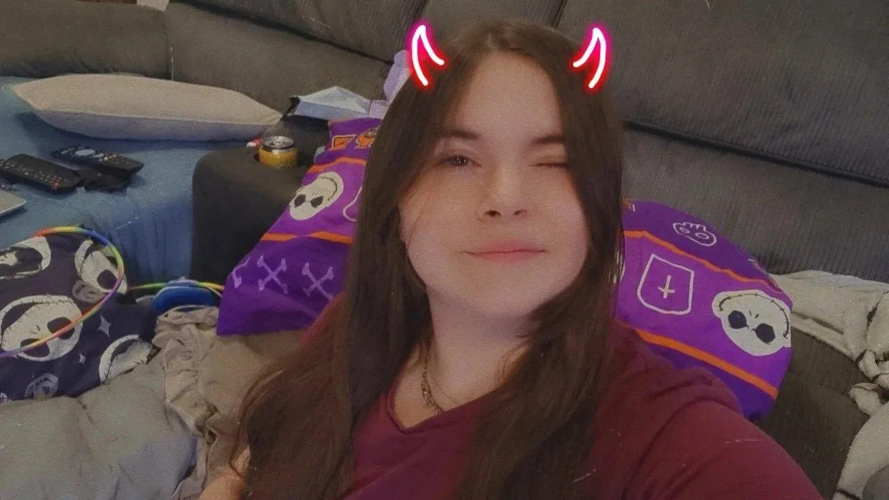 A young woman taking a selfie on a couch, wearing a red shirt, with digital red devil horns and ears added in the photo. She is winking and smiling, with long brown hair, in a cozy room with pillows, a blanket, remote controls, and snacks visible in the background.