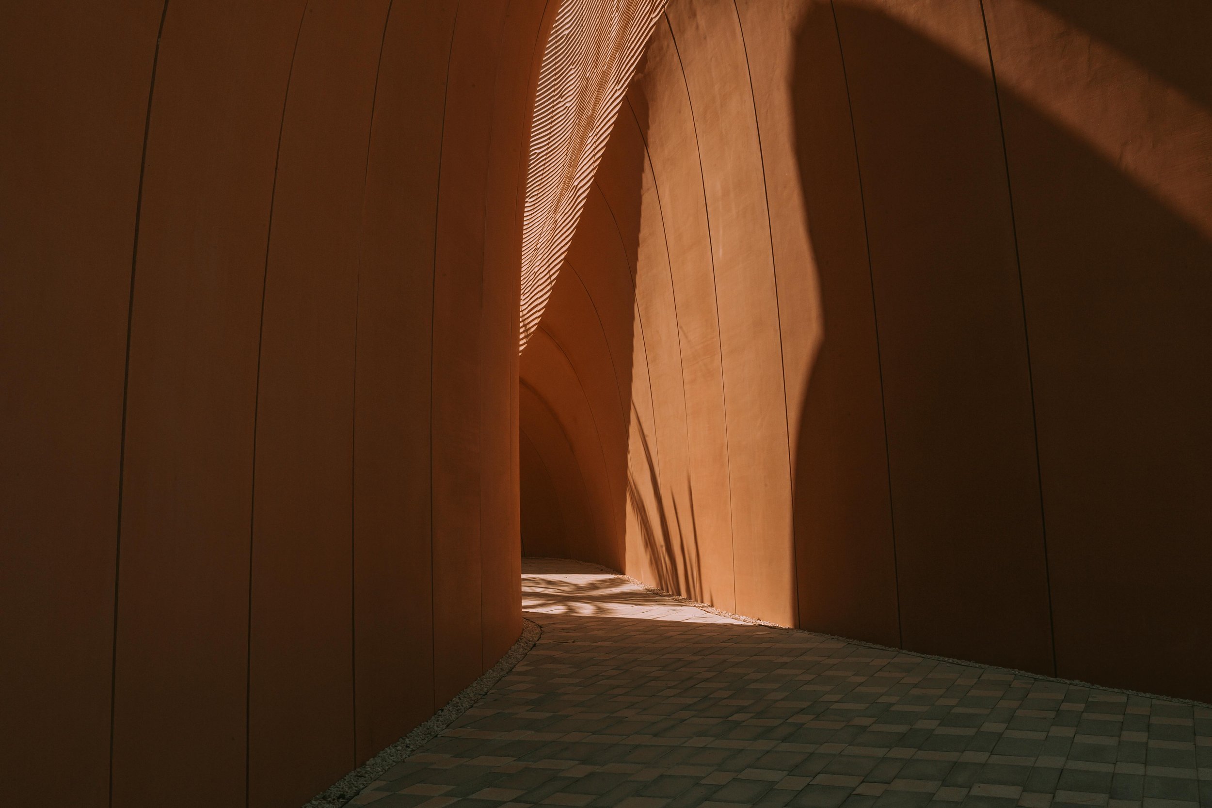 Curved wooden wall with soft lighting and a cobblestone pathway leading through an architectural corridor.