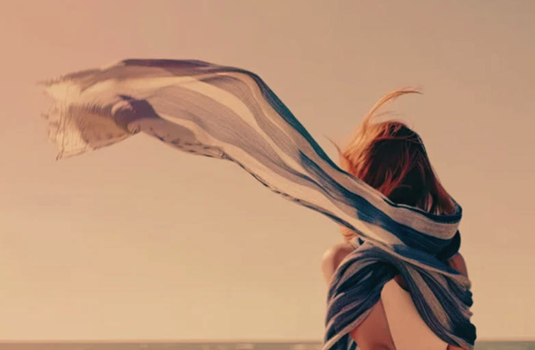 A woman with short hair wrapped in a flowing striped scarf, standing outdoors against a clear sky at sunset.