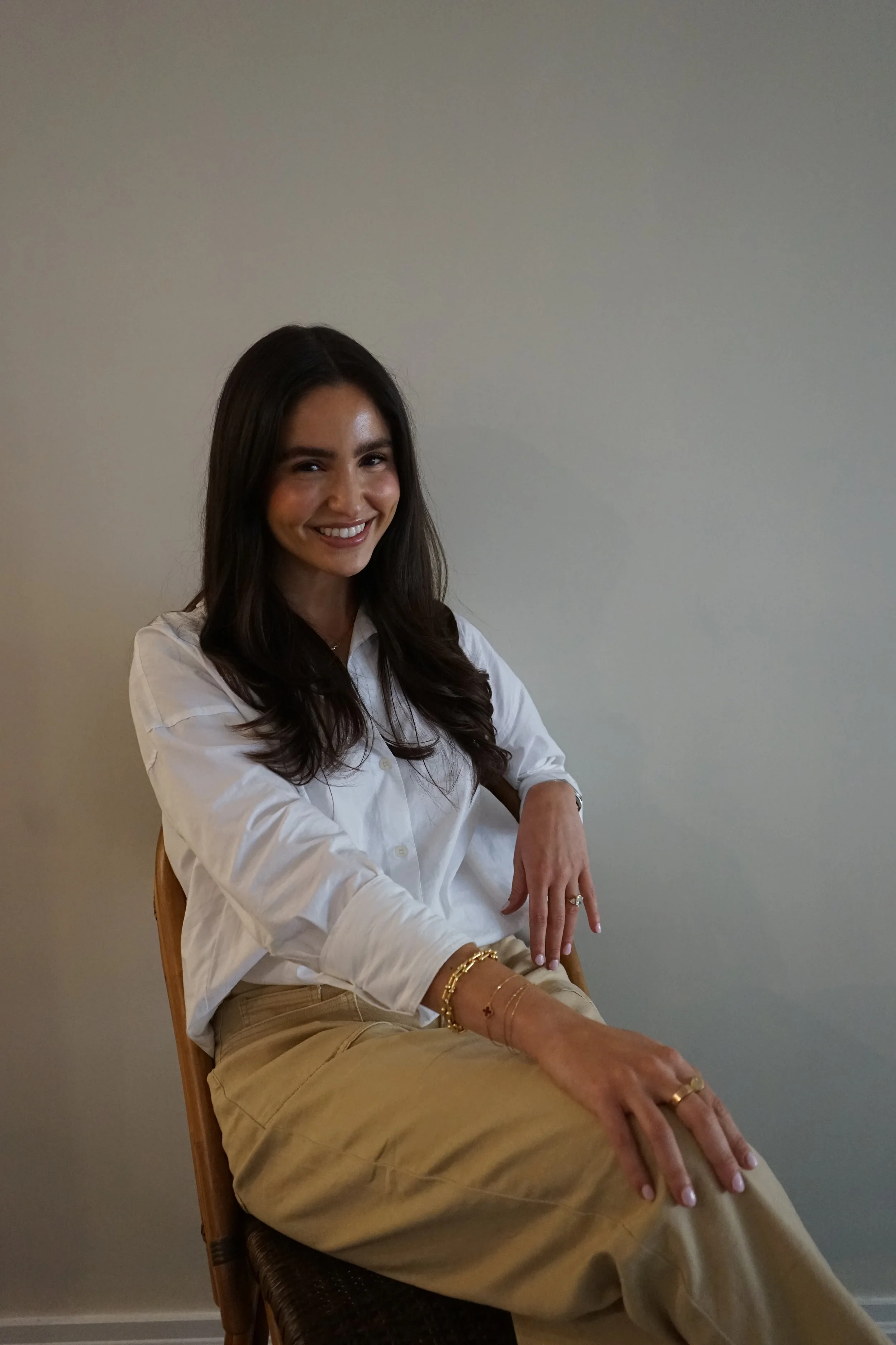 A woman with long dark hair and a white shirt is sitting on a chair against a plain gray wall, smiling at the camera.