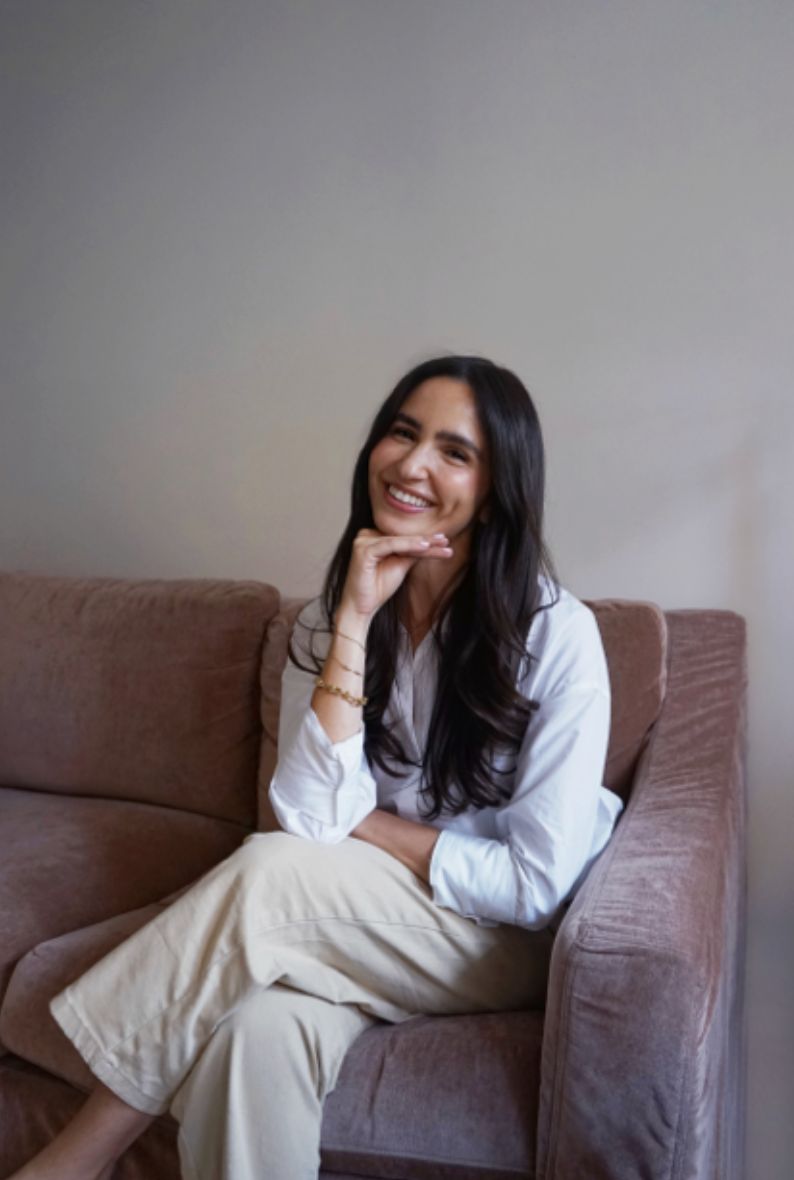 A woman with long dark hair, smiling, sitting on a plush pink couch, wearing a white blouse and beige pants, with her right hand resting under her chin.