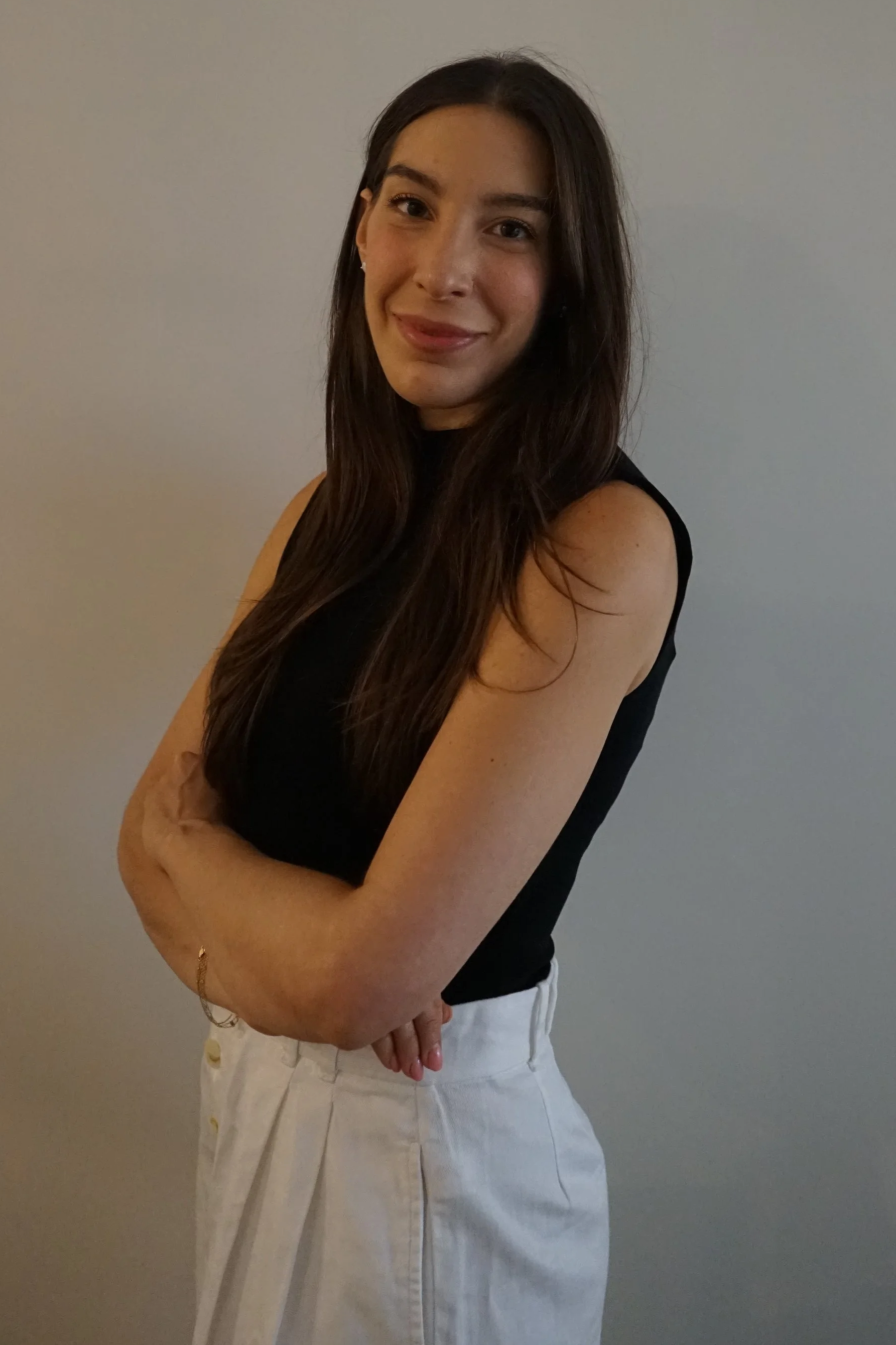 A woman with long brown hair, wearing a sleeveless black top and light-colored pants, standing against a plain gray wall, looking at the camera with a soft smile.
