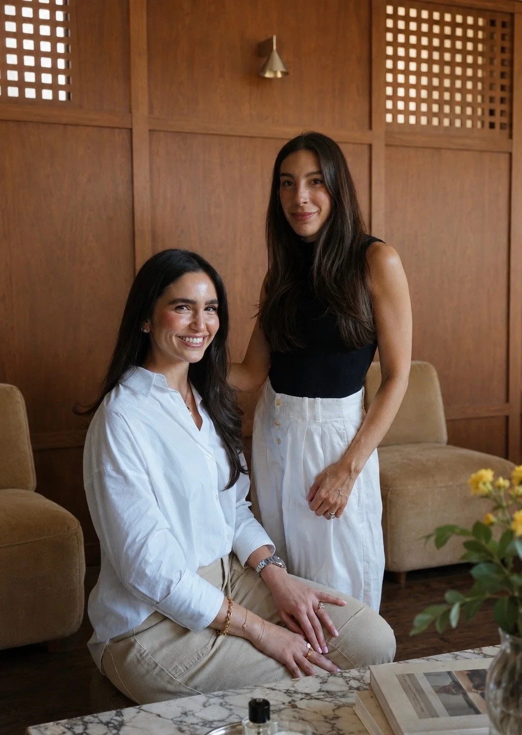 Two women with long dark hair in a room with wooden paneling. One is sitting on a marble-topped table, wearing a white blouse and beige pants, with gold jewelry. The other is standing, wearing a black sleeveless top and white pants. They are smiling and pose together.