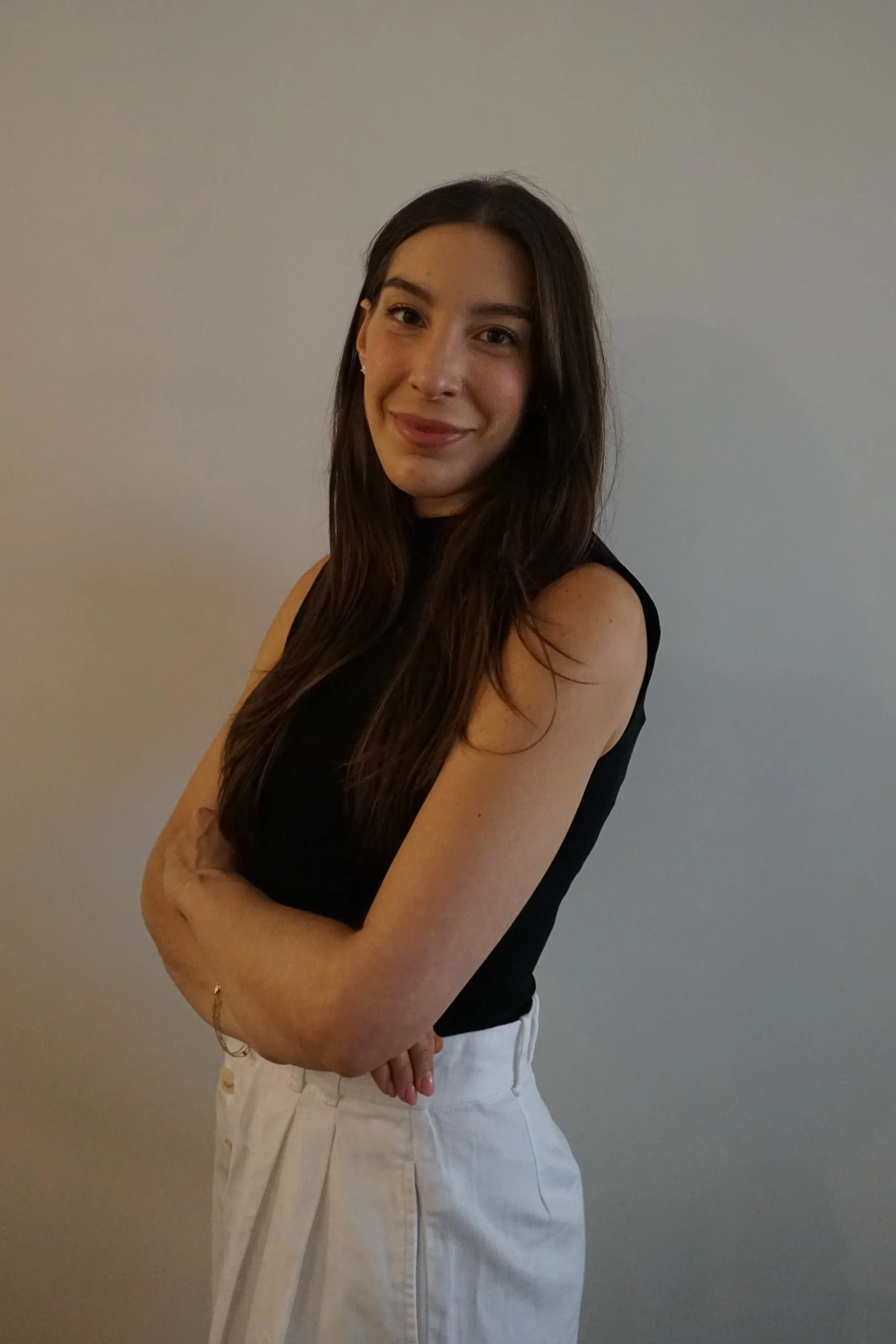 A young woman with long brown hair, wearing a black sleeveless top and white pants, standing with crossed arms against a plain gray wall.
