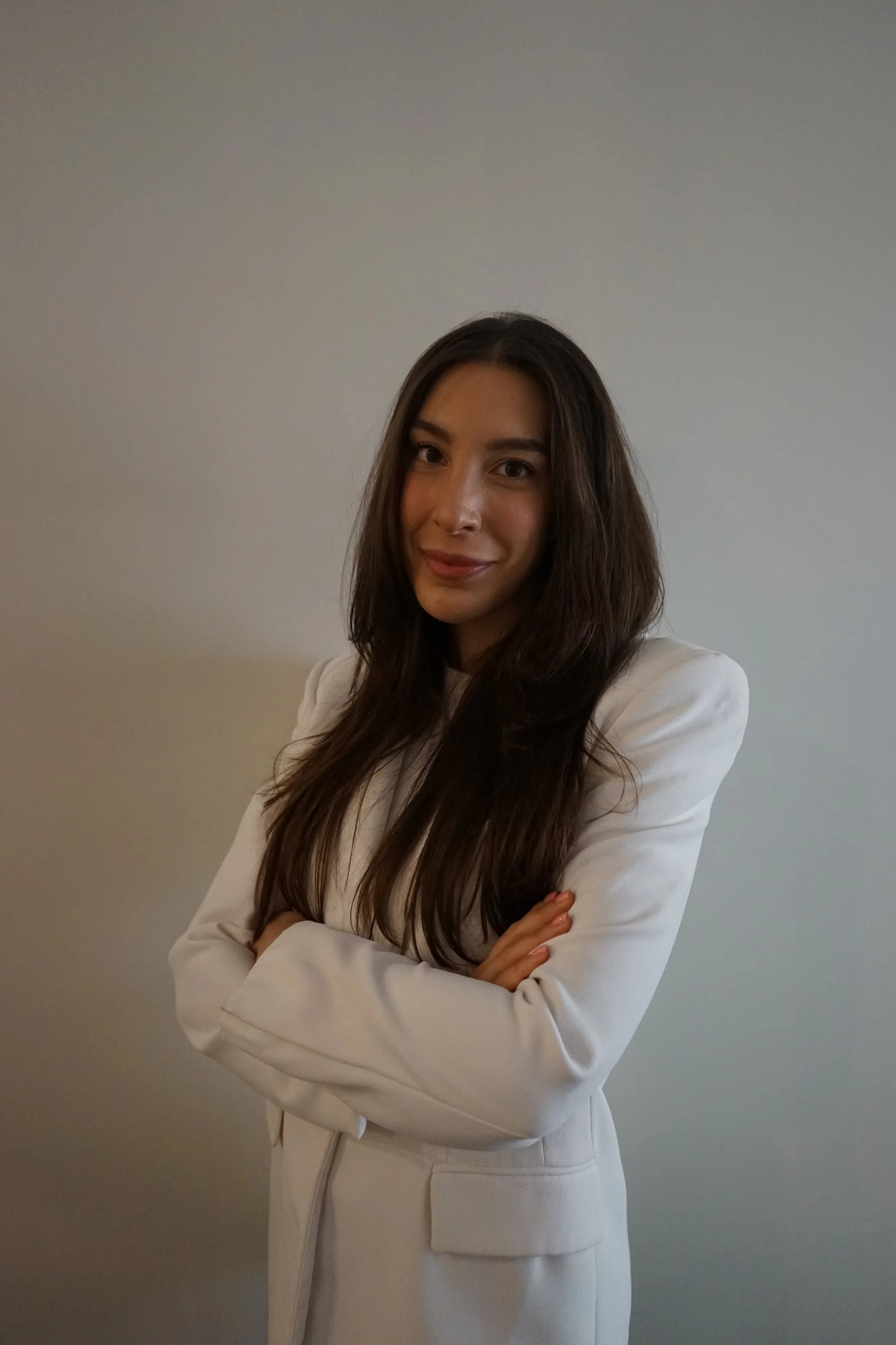A woman with long brown hair, dressed in a white blazer and skirt, standing against a plain light-colored wall with her arms crossed, smiling softly at the camera.