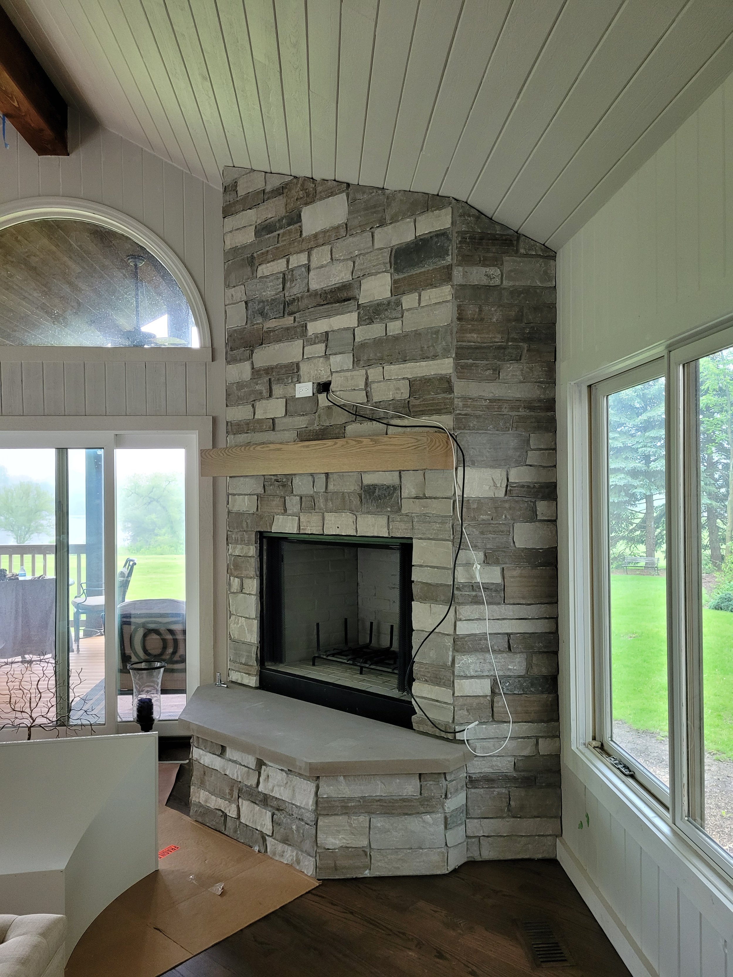 A living room features a stone fireplace, large windows with white frames, and a view of a green outdoor space. There is a sliding glass door leading outside, and a partial view of a ceiling fan can be seen through the window. The ceiling is paneled, and there is a piece of wood and some electrical wiring attached to the fireplace.