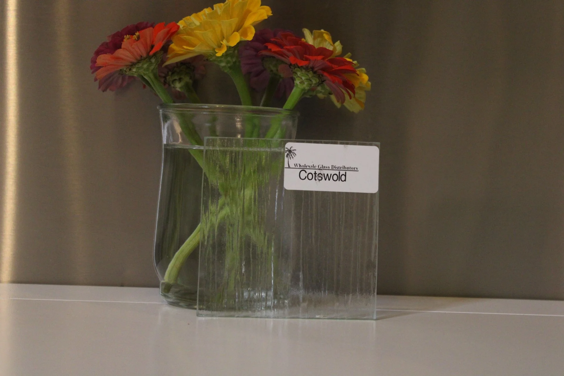 Colorful Gerbera daisies in a glass vase on a shelf, with a label from Cotswold wholesale glass distributors, against a plain background.