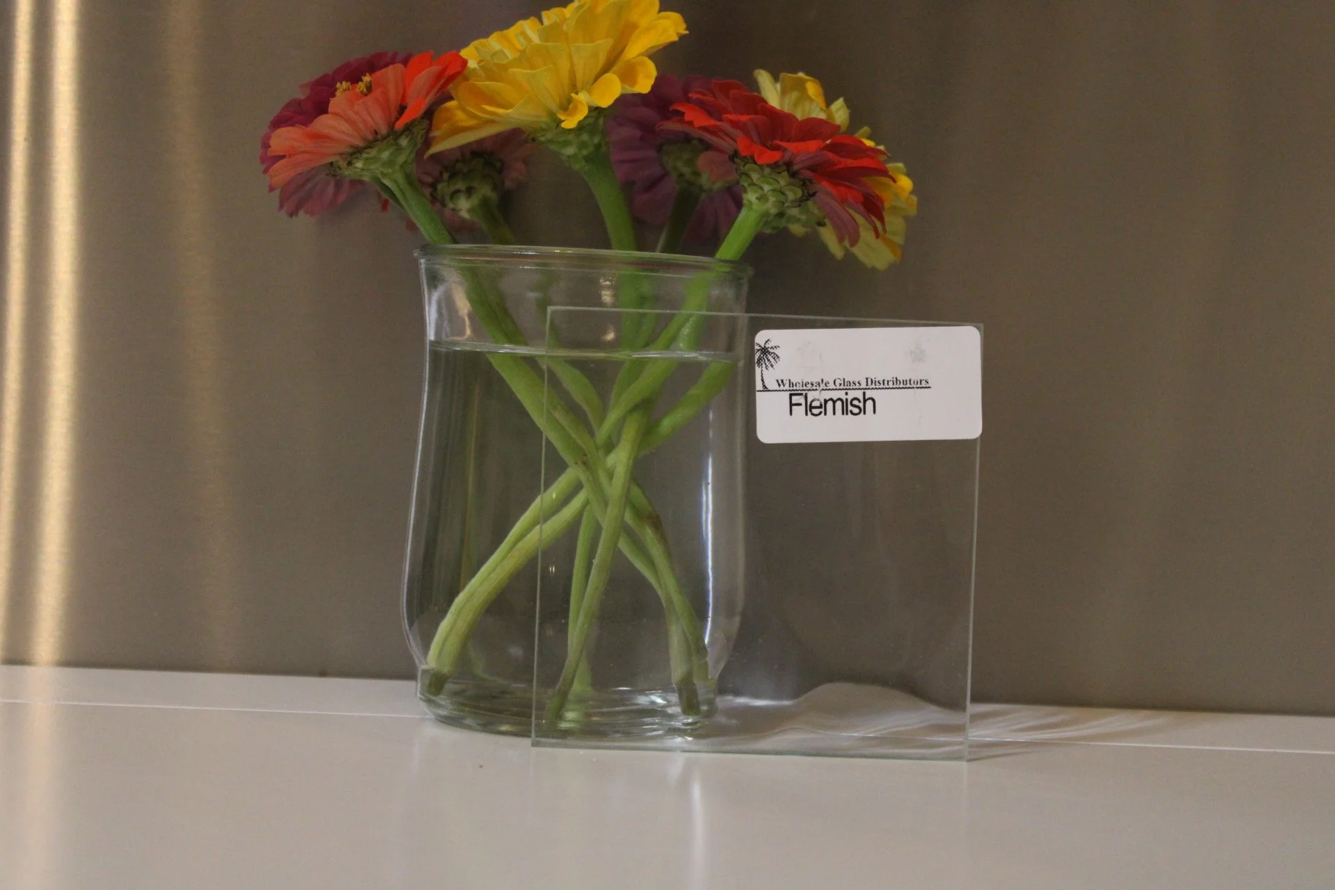A bouquet of colorful flowers with pink, yellow, orange, and purple petals in a clear glass vase filled with water, placed on a white surface against a dark gray or metallic background. There is a small white label on the vase that reads 'Flemish' an