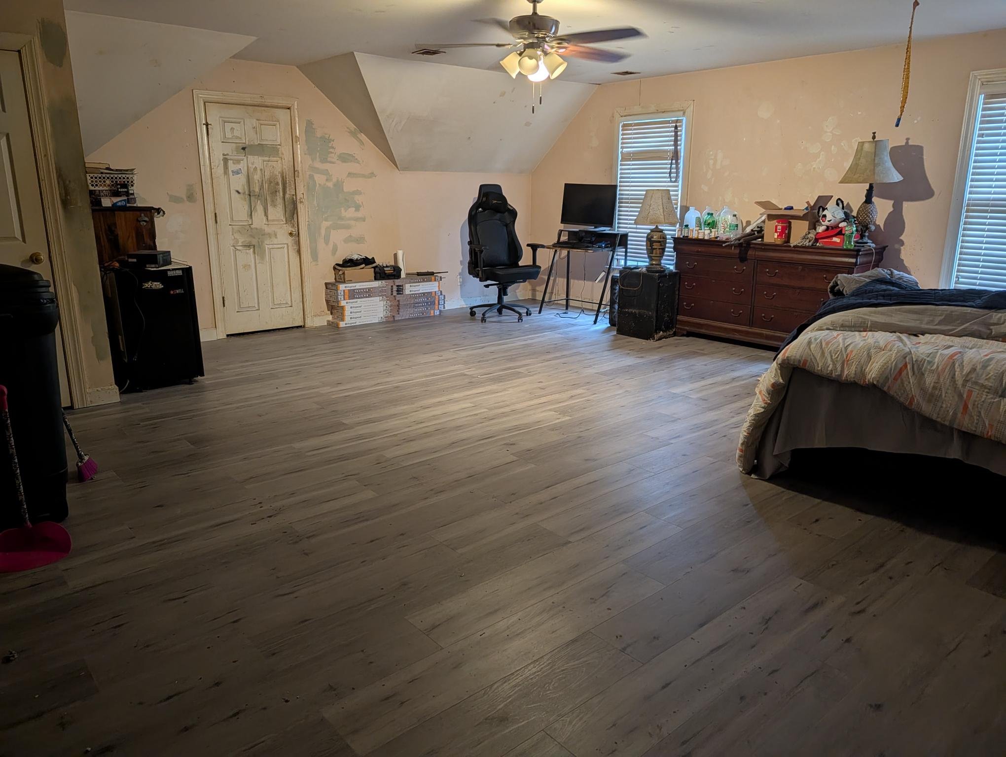 Spacious bedroom with wooden floors, two windows with blinds, an unmade bed on the right, a large dresser with lamps and various items, a black office chair, a desk with a computer, and some boxes near a door with peeling paint.