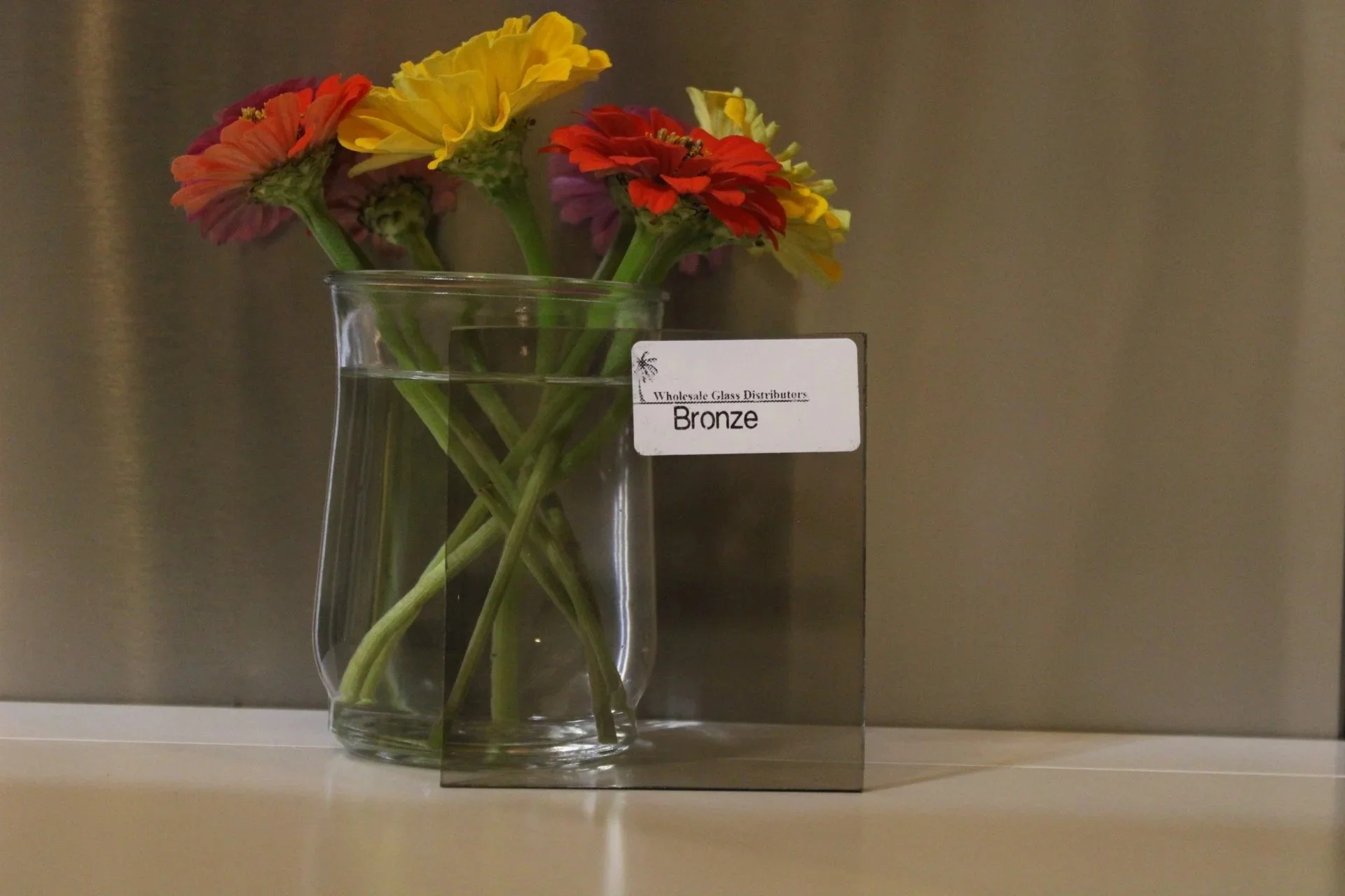 A clear glass vase holding a bouquet of colorful flowers, including yellow, red, and pink blooms, placed on a light-colored surface against a neutral background, with a small white label on the vase reading 'Wholesale Glass Distributors Bronze'.