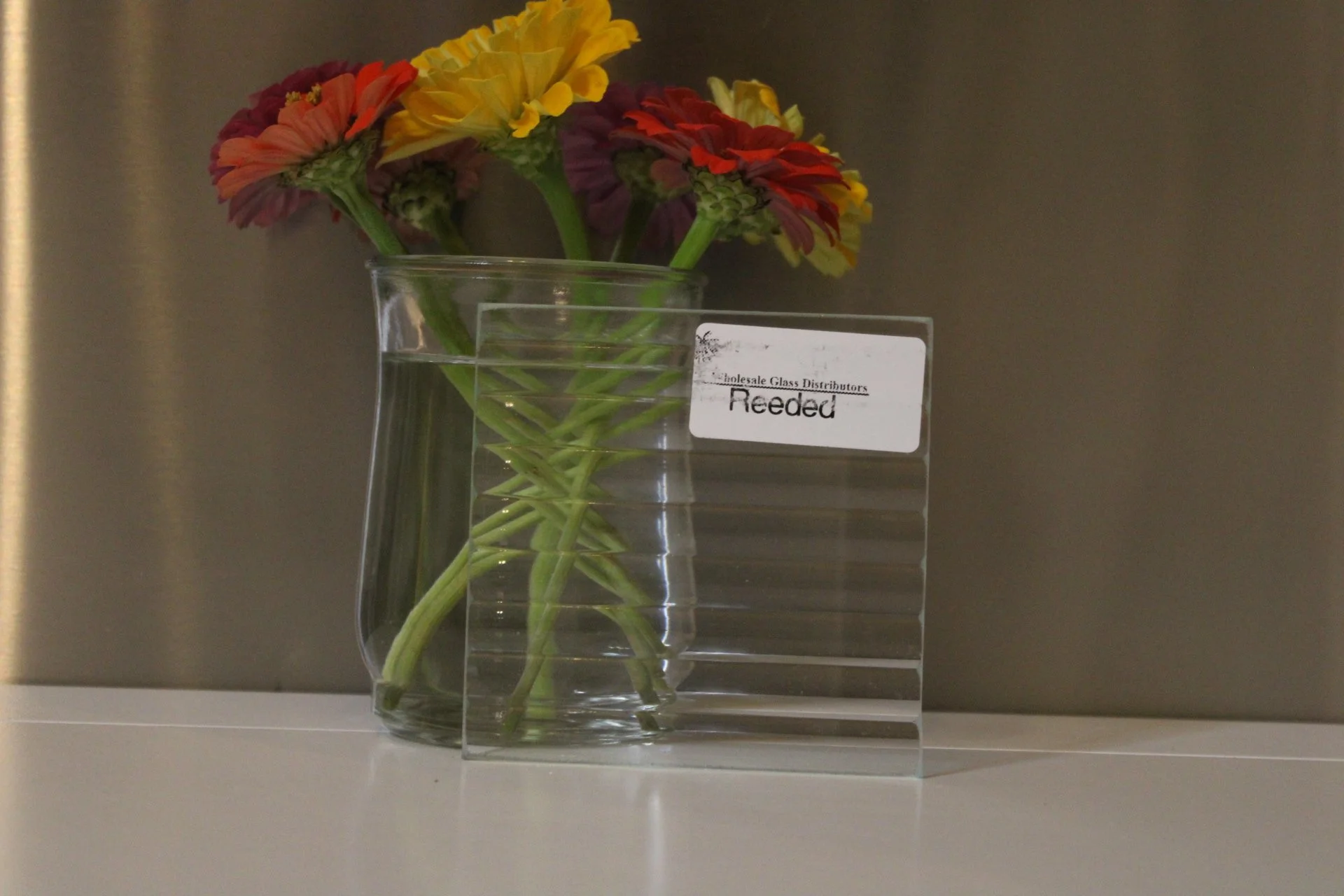 A clear glass vase with colorful flowers, including red, yellow, and purple blooms, placed on a white surface against a grey background. A label on the vase reads 'Reeded'.