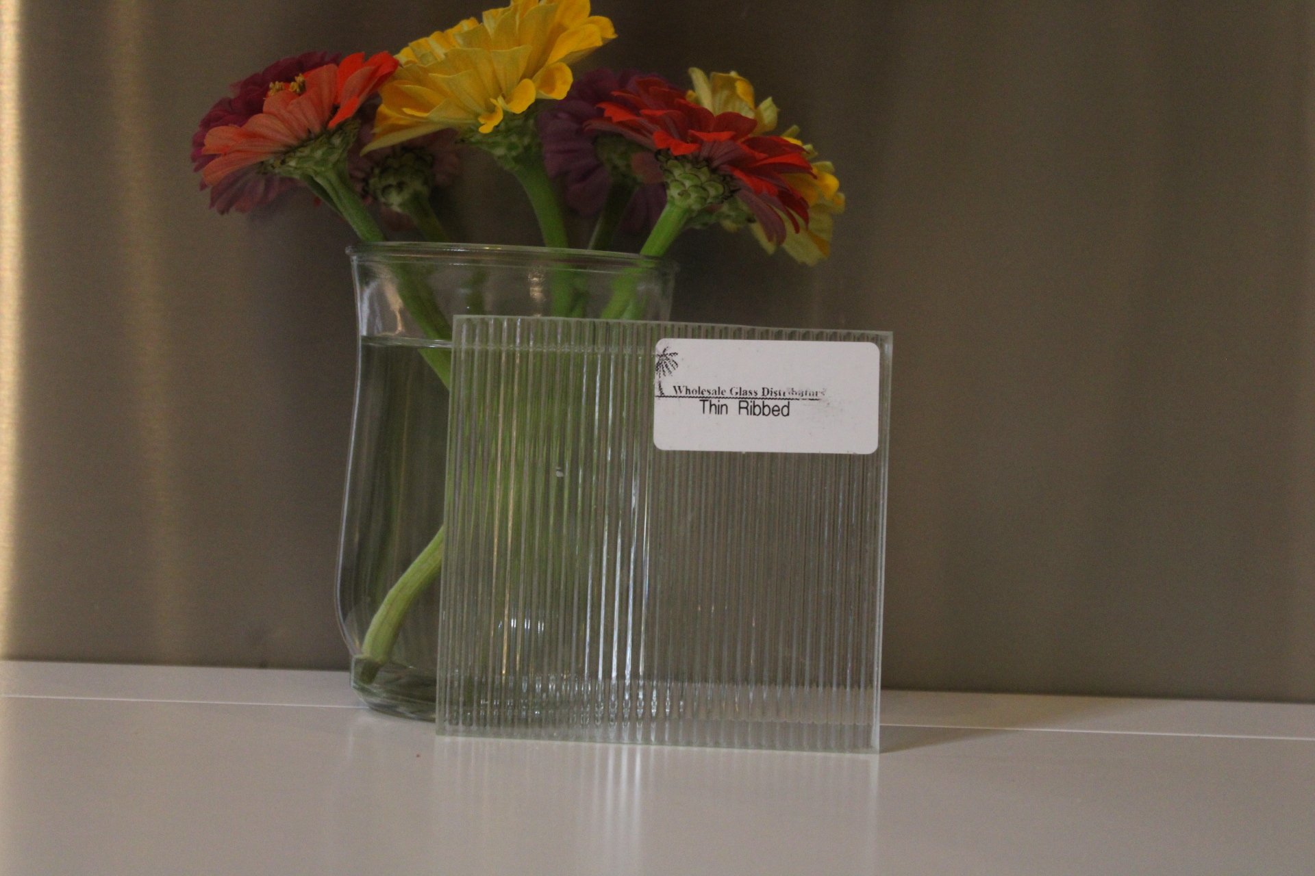 A clear glass vase holding orange, yellow, and purple flowers, with a second ribbed glass container with a white price tag labeled 'Thin Ribbed' positioned in front.