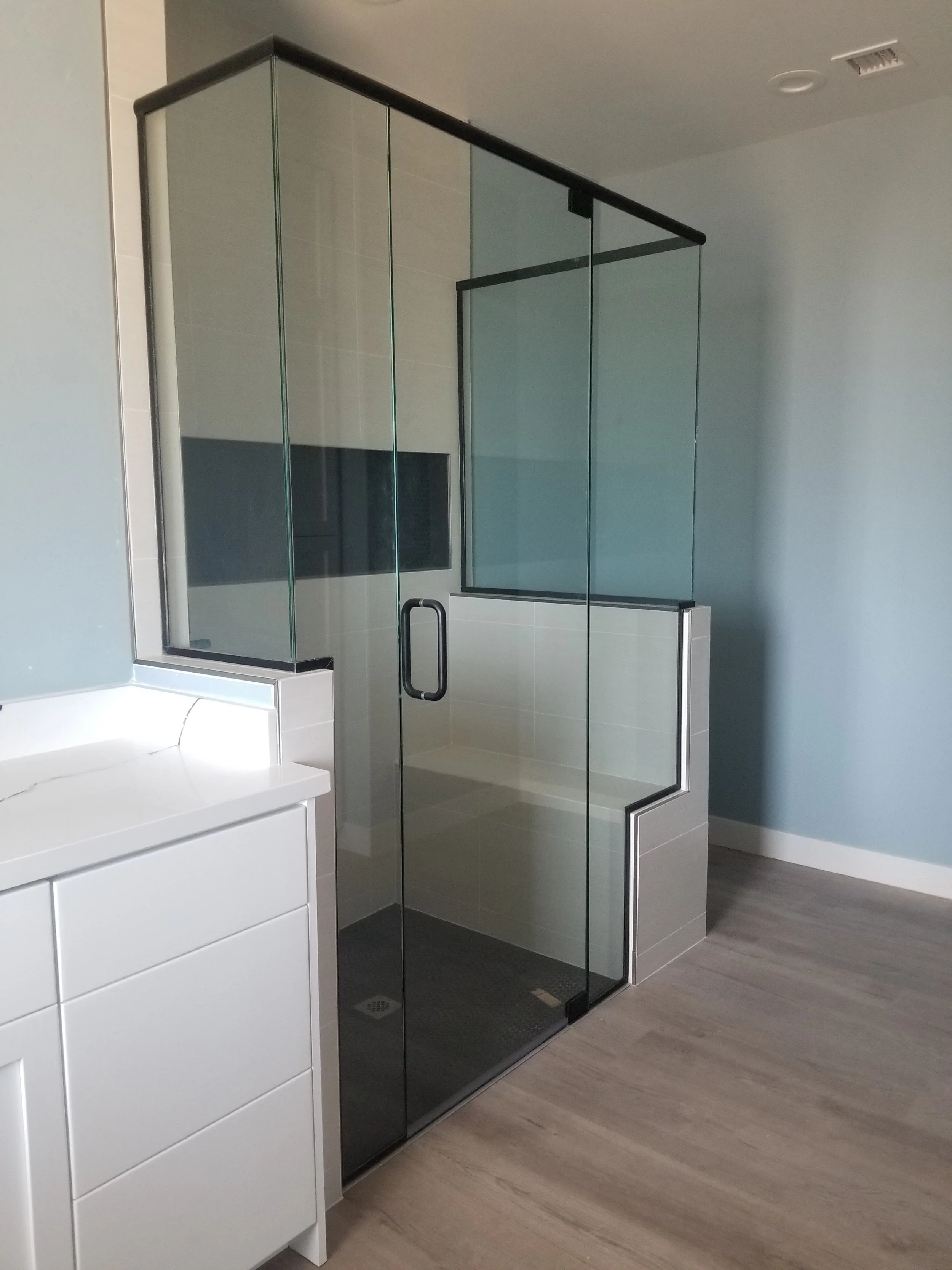 A modern bathroom shower with black-framed glass walls next to a white vanity with drawers and a white top, set on a wood-tone floor with light blue walls.