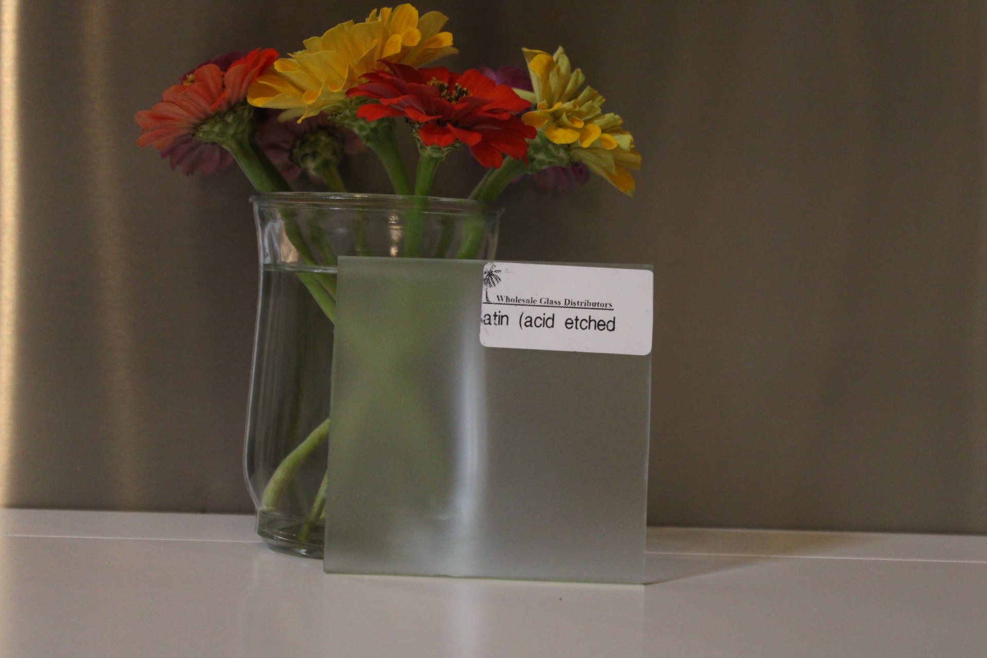 A clear glass vase with colorful flowers, including yellow, orange, and red blossoms, on a table against a plain background, with a white label attached to the vase.