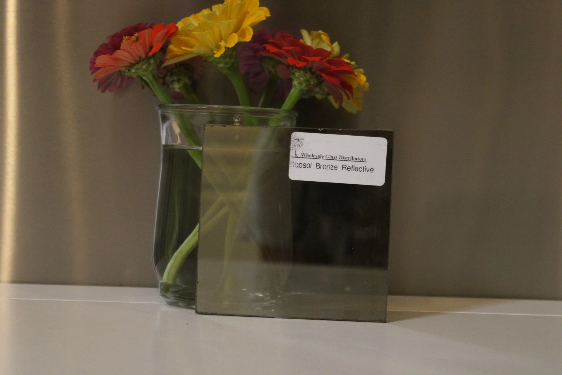 A clear glass vase with yellow, red, and purple flowers and green stems inside, placed on a white surface. There is a black informational card partially visible next to the vase, with the words 'Wholesale Glass Distributors' and 'Topsol Bronze Reflec