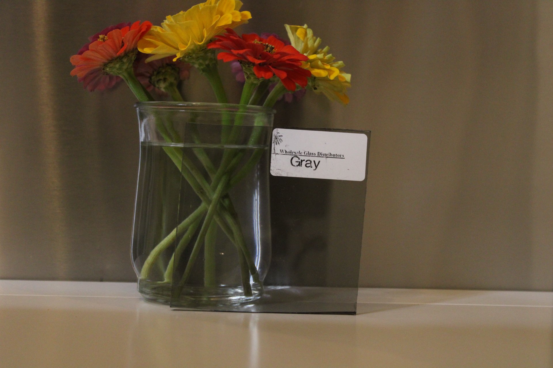 A glass vase with water holding a bouquet of multicolored flowers, including yellow, red, and pink, placed on a shelf against a neutral background with a white label reading 'Wholesale Glass Distributors Gray'.