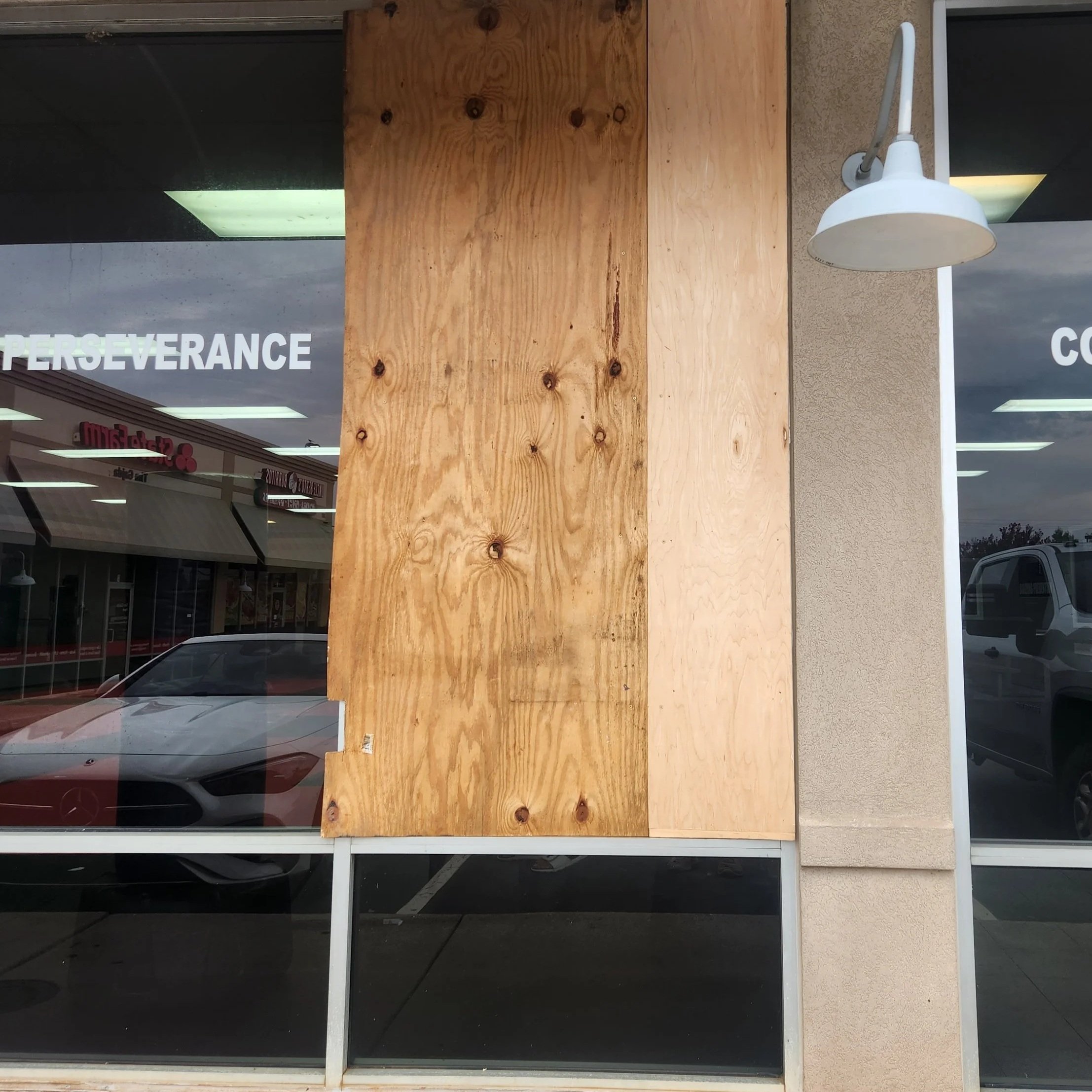 Large piece of plywood attached to glass window or door of a commercial building, with the word 'PERSEVERANCE' partially visible on the glass. The plywood covers most of the window, and a white outdoor light fixture is mounted on the building adjacent to the plywood.