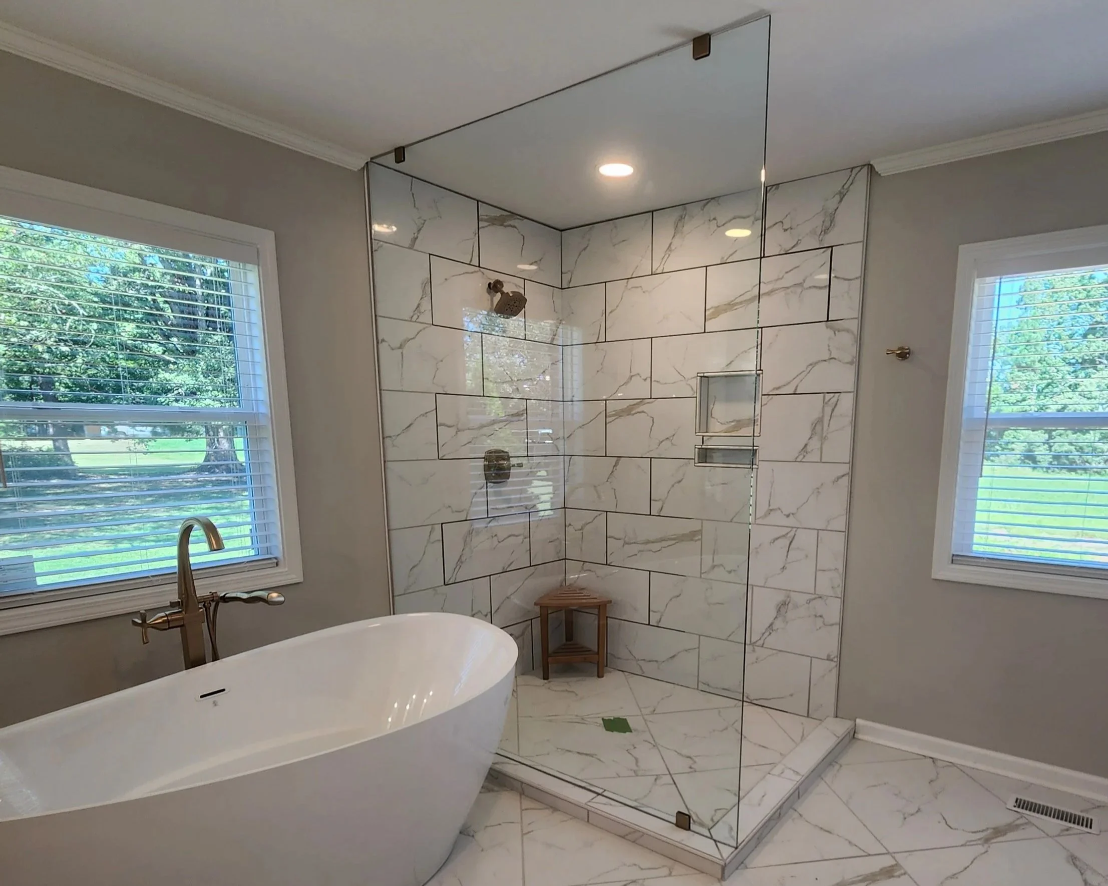 Bathroom with white freestanding bathtub, marble-tile shower with glass door, window with blinds, and small wooden stool.